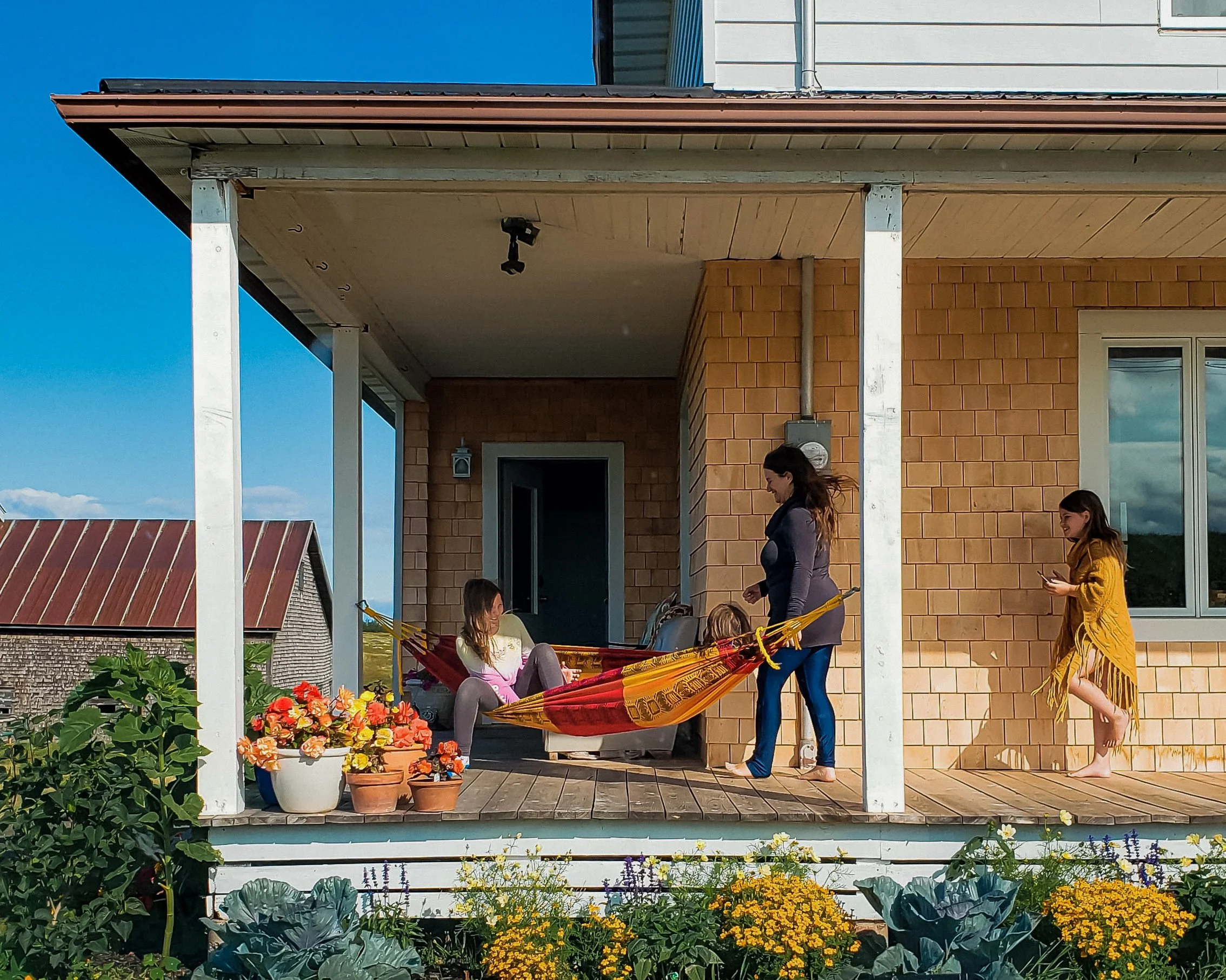 A group of people standing and sitting on a porch with a beige brick wall, white pillars, and a wooden deck, surrounded by potted flowers and plants under a blue sky.