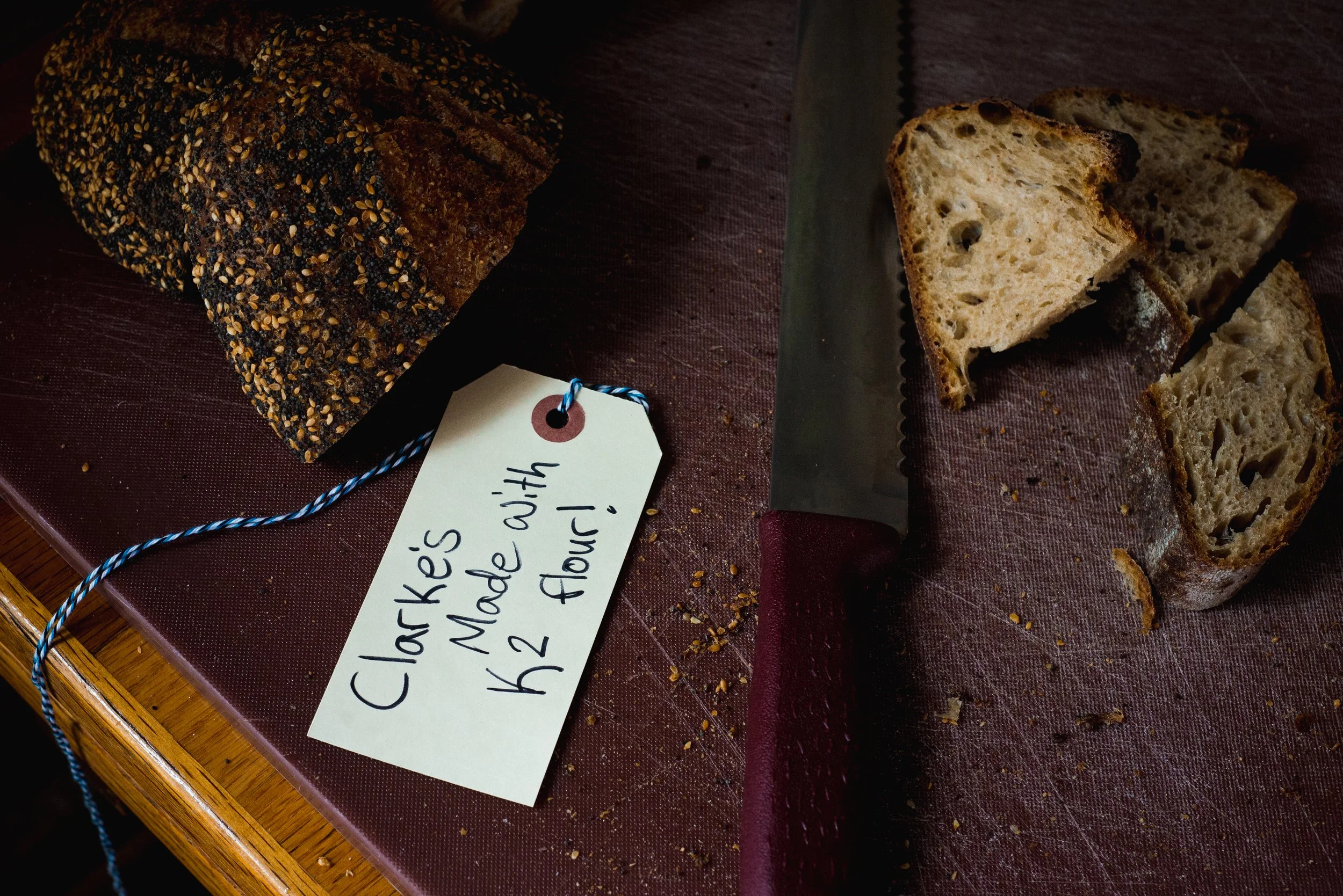 Sourdough bread on a cutting board with a loaf on the left covered in seeds and several slices on the right showing a airy crumb. A bread knife is also on the board. A handwritten tag reads 'Clarke's Made with K2 Flour!'
