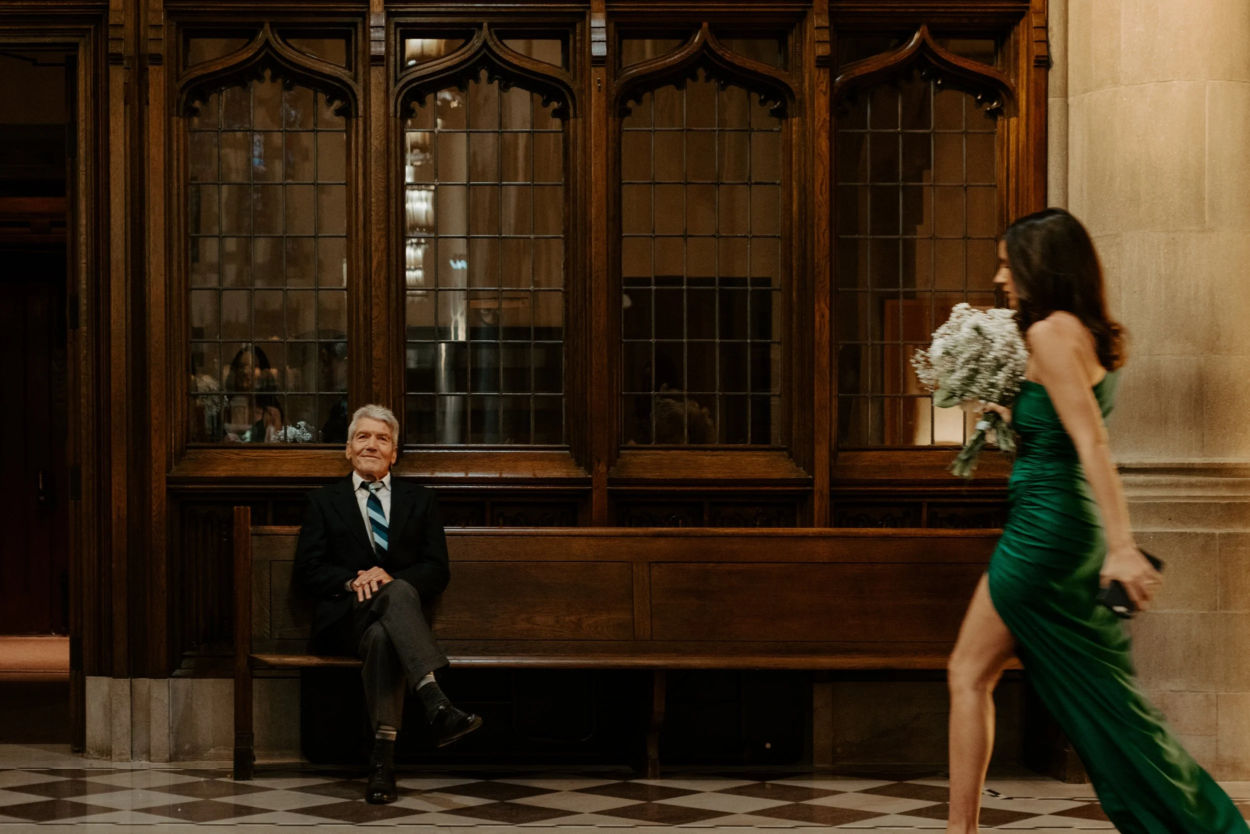 Father of the bride sitting on a Montreal church pew, smiling at the camera with crossed legs, as a bridesmaid with a bouquet walks past in motion blur.