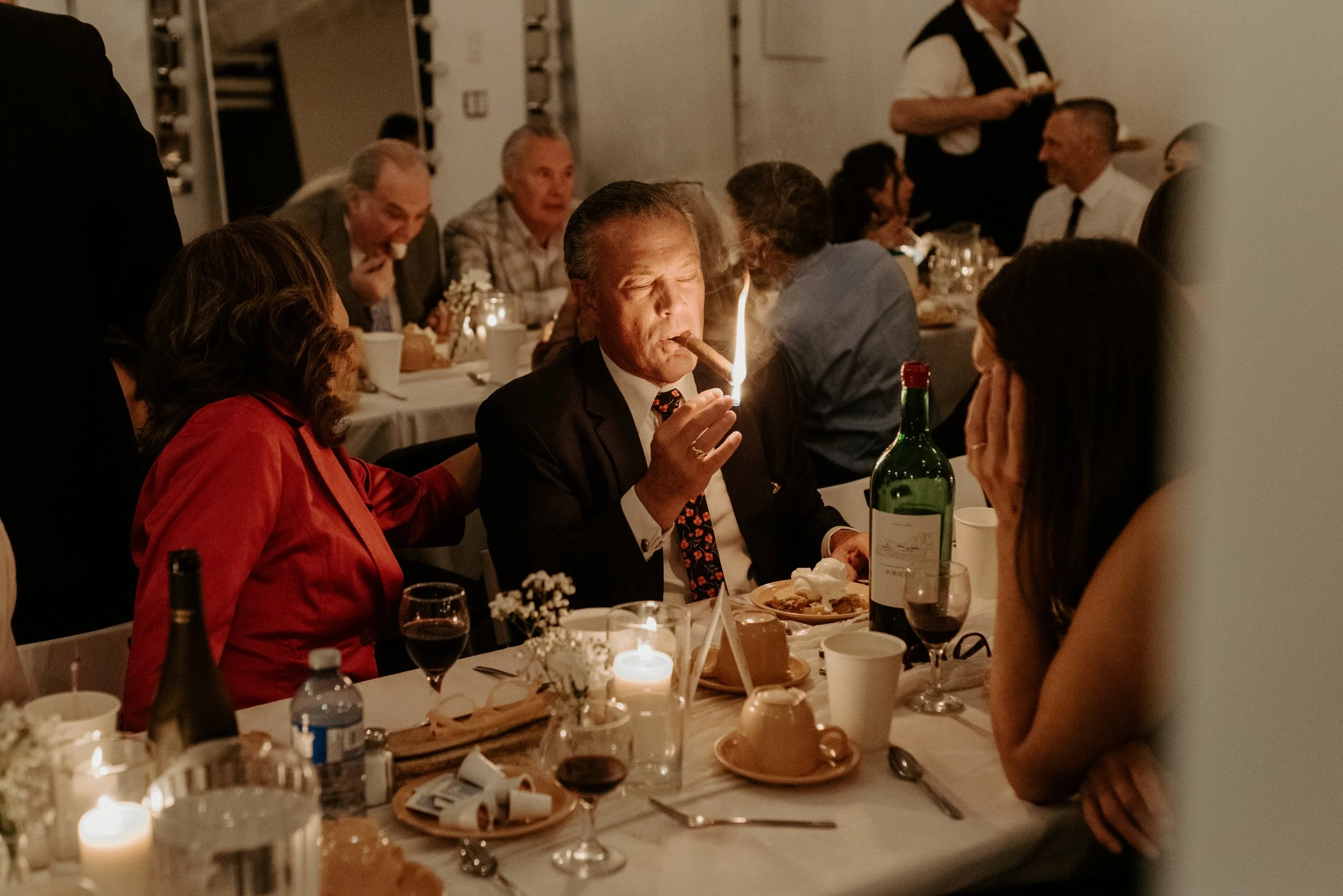 Candid wedding photo of the groom's father lighting a cigar at a dimly lit reception table surrounded by socializing guests.