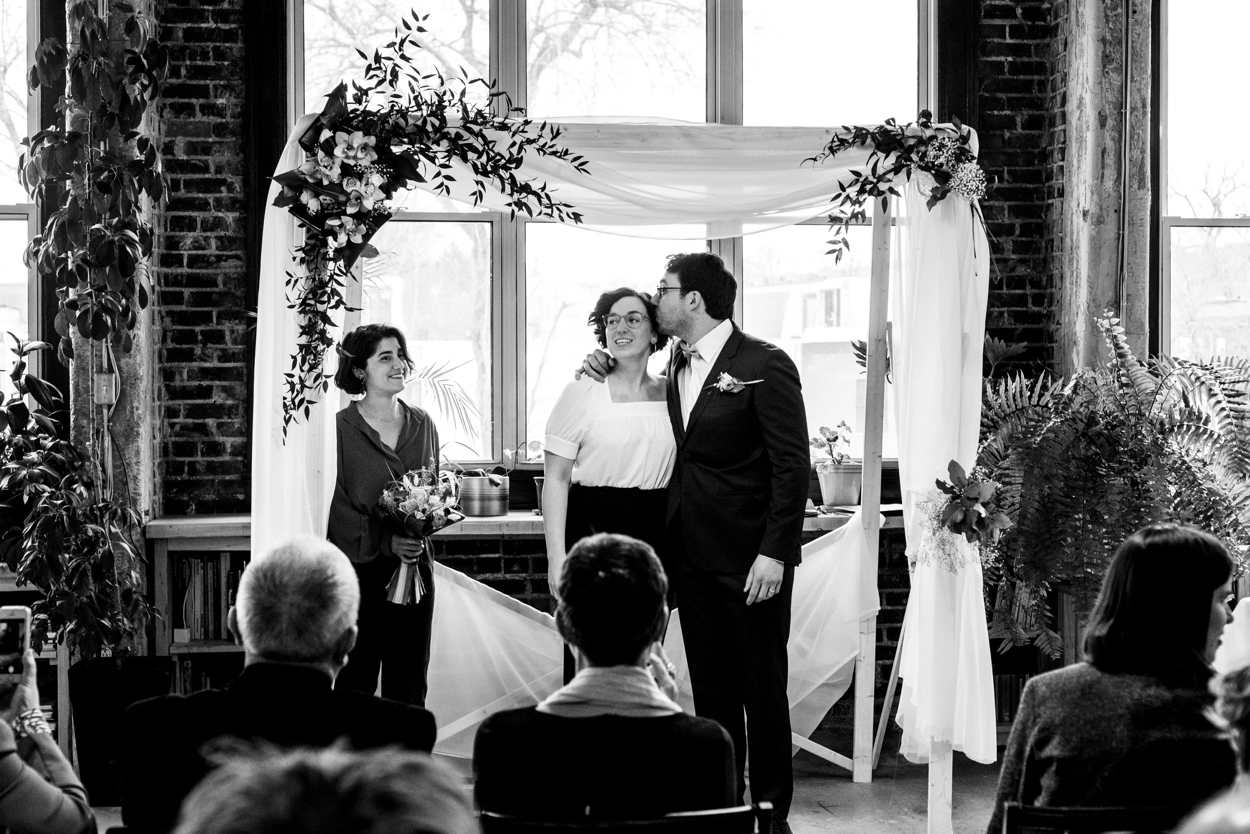 Black and white portrait of groom kissing forehead of bride in front of altar as celebrant watches on. 