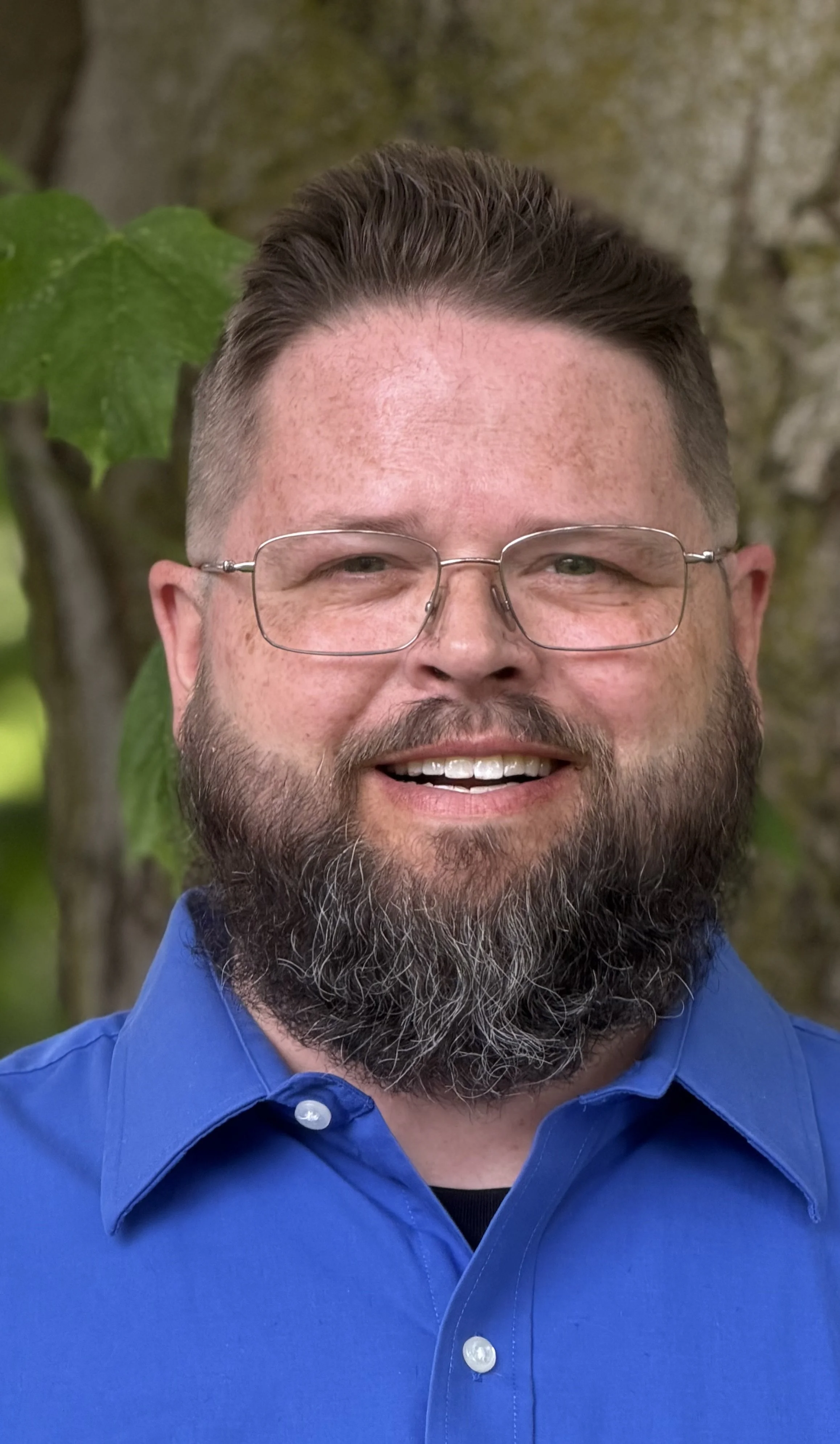 Close-up of a smiling man with glasses, a beard, and short hair, wearing a blue collared shirt, outdoors with trees and green leaves in the background.