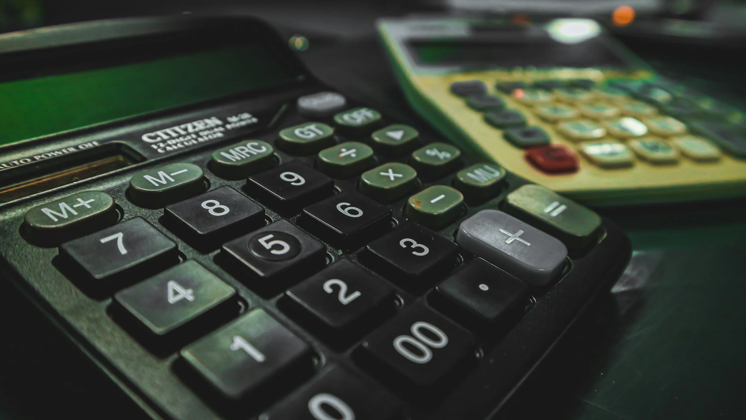Close-up of a calculator with black and gray buttons on a dark surface, with another digital device partially visible in the background.