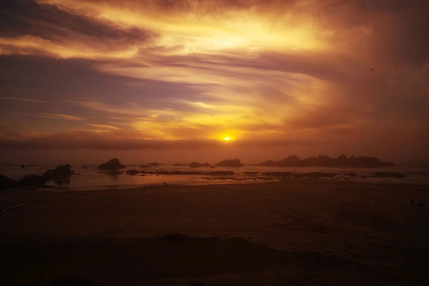 Sunset over a beach with rocks in the ocean and a few people walking on the sand.