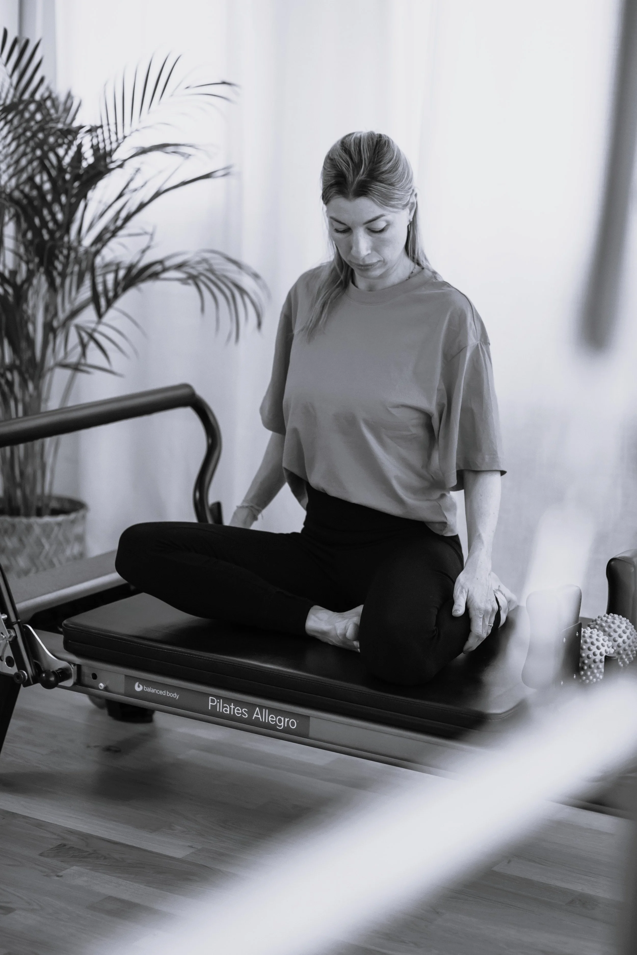 Frau sitzt auf einem Pilates-Gerät, macht eine Dehnübung, im Hintergrund ist eine Pflanze.