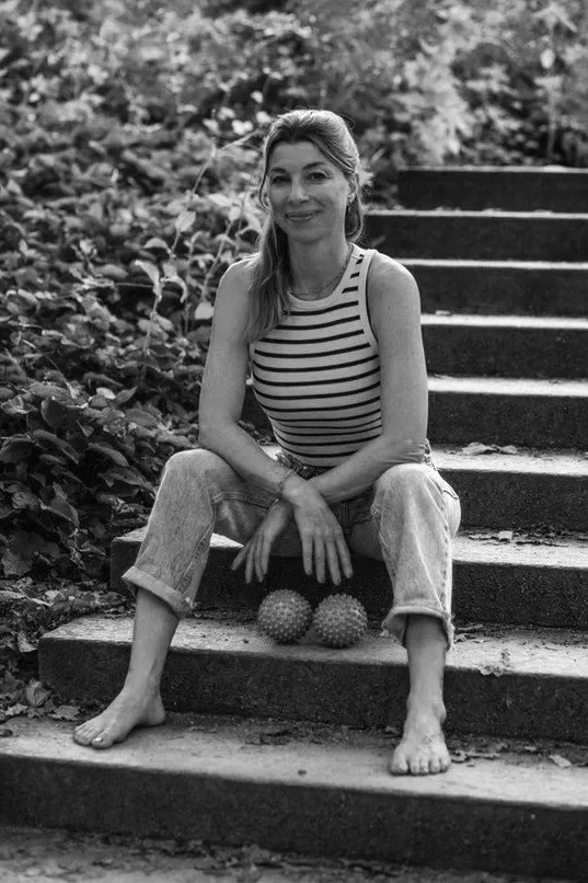 Eine Frau im gestreiften Top und Jeans sitzt barfuß auf einer Treppe im Freien, umgeben von Natur, mit zwei spikigen Bällen vor ihr.