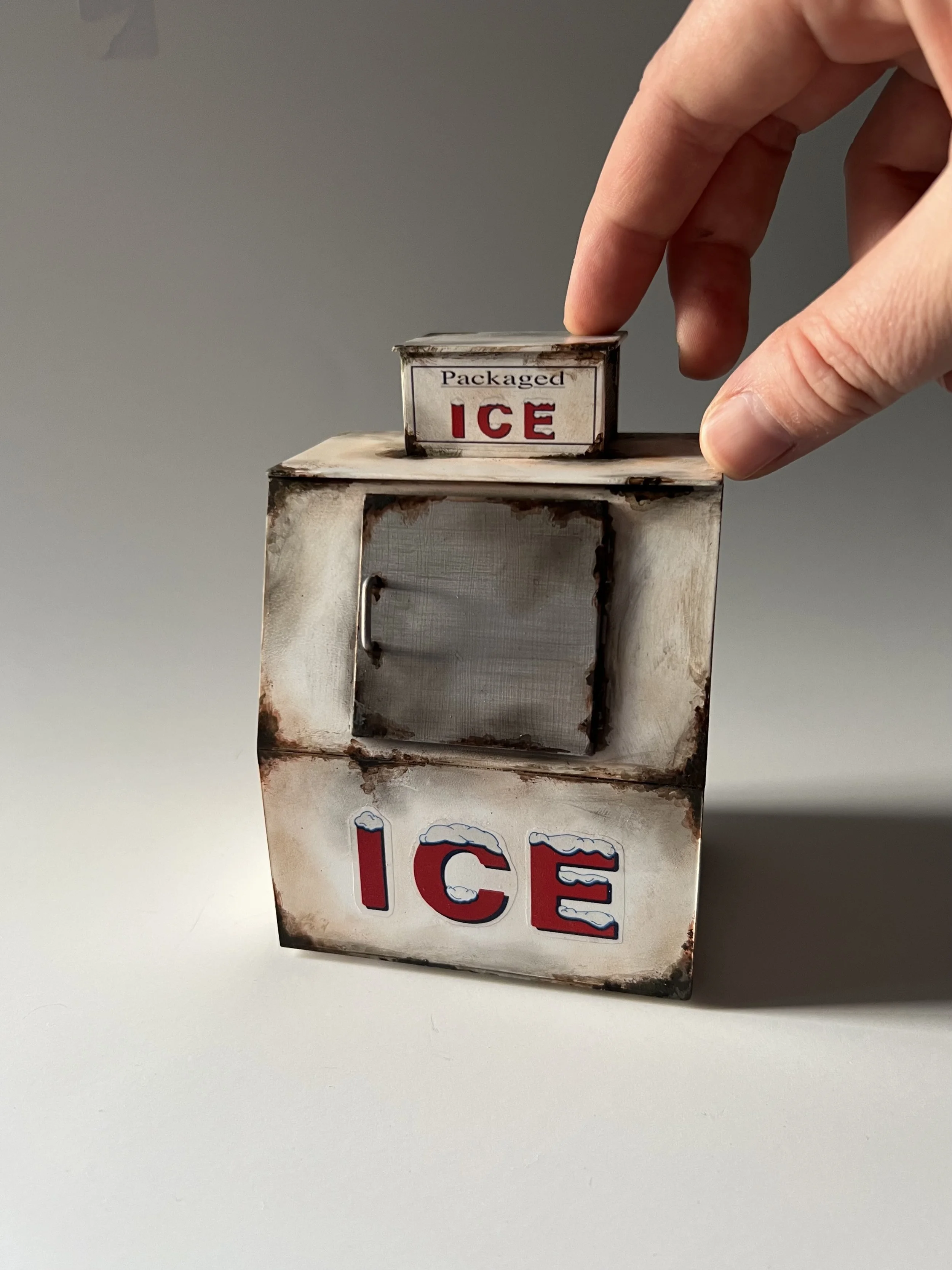A hand adjusting the lid of a small vintage metal ice box with a partially visible 'ICE' label on the side.