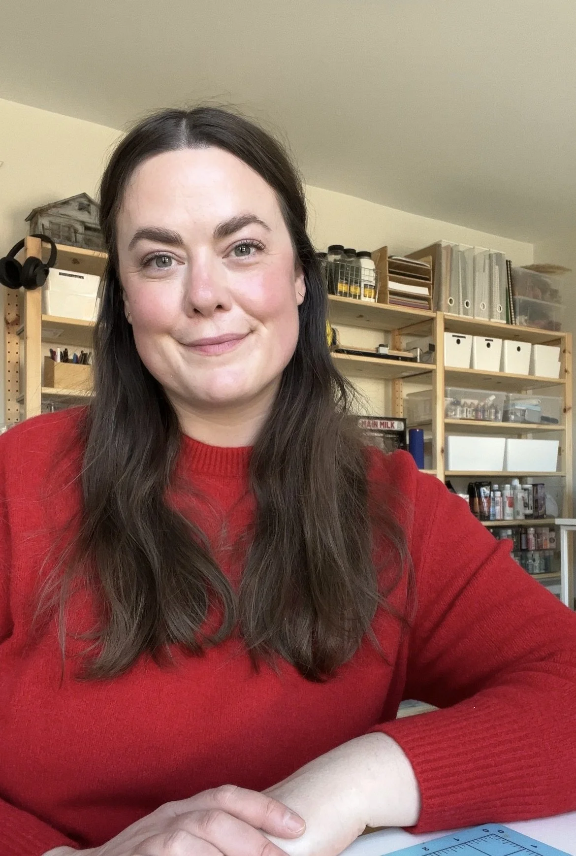 A woman with long brown hair and gray eyes wearing a red sweater, smiling and sitting at a table in a room with wooden shelves filled with books, binders, and craft supplies.