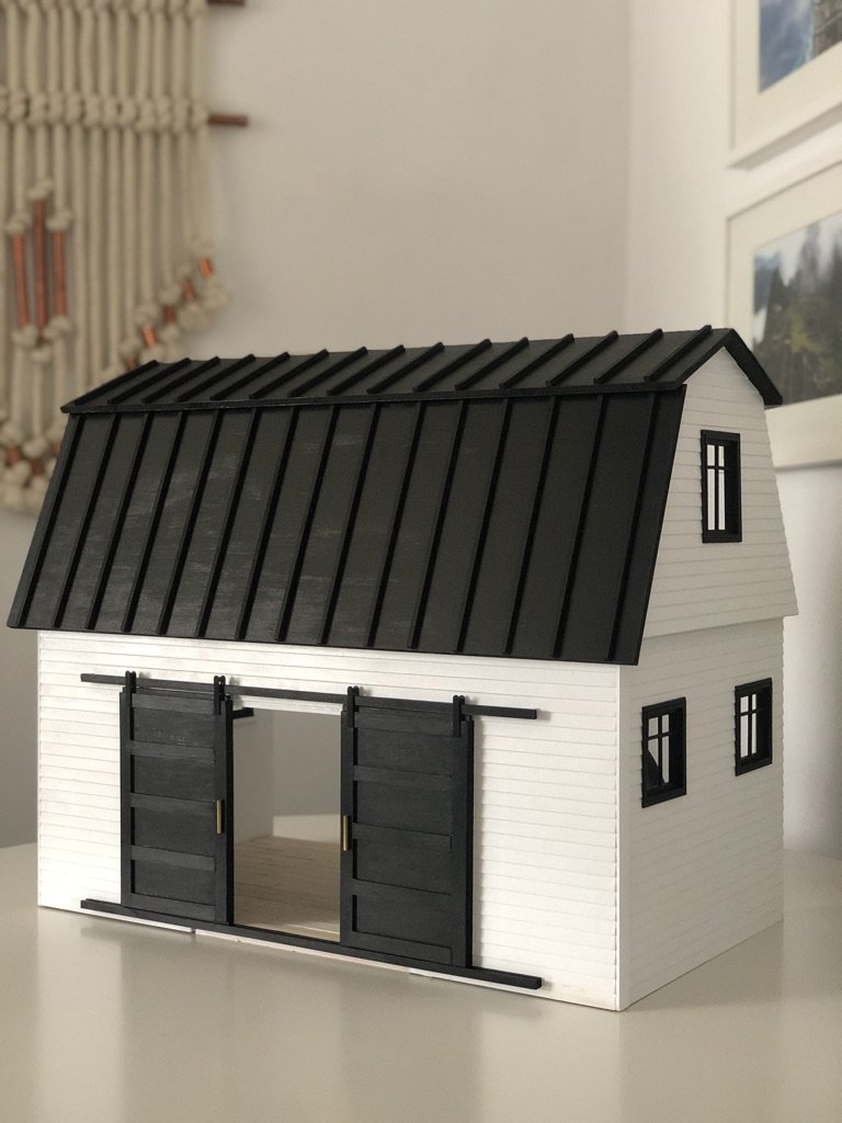 A miniature model of a barn with white siding, black sliding doors, a black roof, and windows, placed indoors on a white surface.