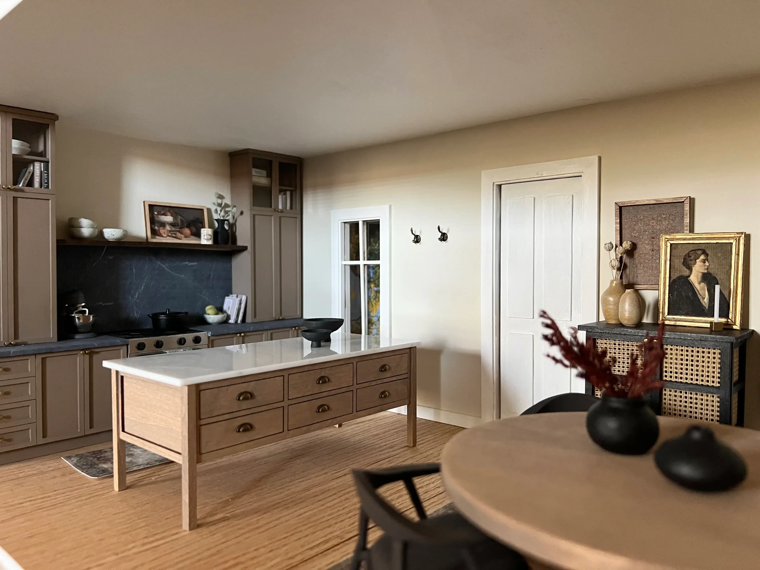 A modern kitchen with beige cabinets, a wooden island with multiple drawers, and decorative items on the shelves. There is a painting and two vases on a side table, with some artwork inside a framed picture. A dining table with a bowl of flowers is in the foreground, and a window with white trim is on the far wall.