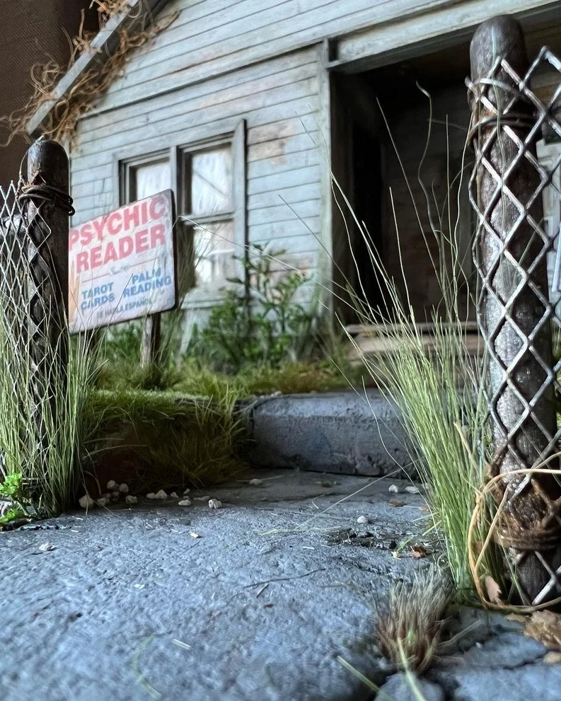A weathered wooden house with a small window, a dark open doorway, and an outdoor area with overgrown grass, weeds, and a weathered sign that reads 'Psychic Reader Tarot Cards Palm Reading.'
