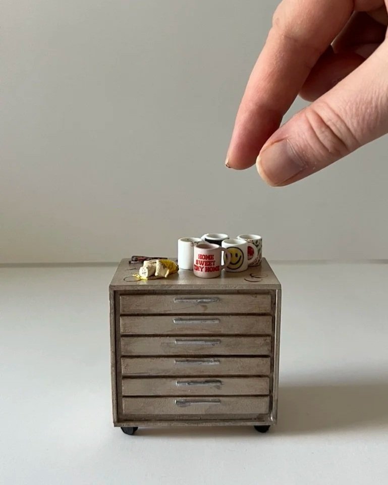 Close-up of a human hand with two fingers, appearing to be pinching or holding, above a miniature wooden filing cabinet with tiny coffee mugs, a small yellow object, and a key on top.