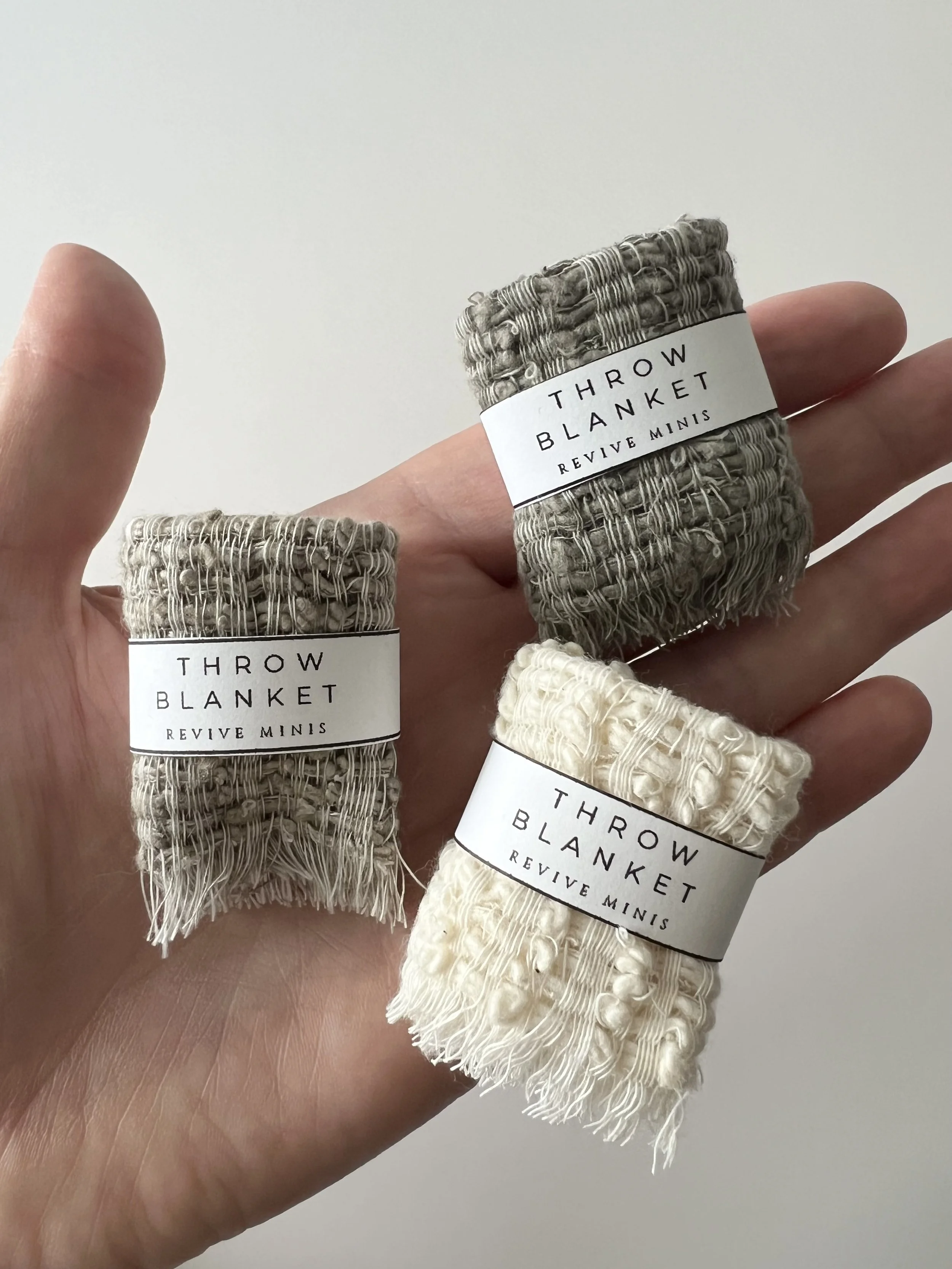Hand holding three rolled-up throw blankets with labels, one in black, one in beige, and one in cream, all labeled "Throw Blanket Revive Minis."