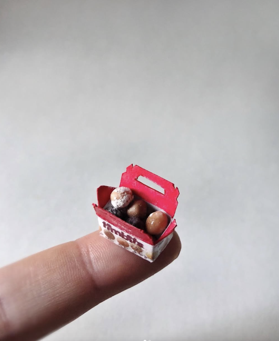 Miniature red shopping basket filled with tiny chocolate candies, held on a person's finger.