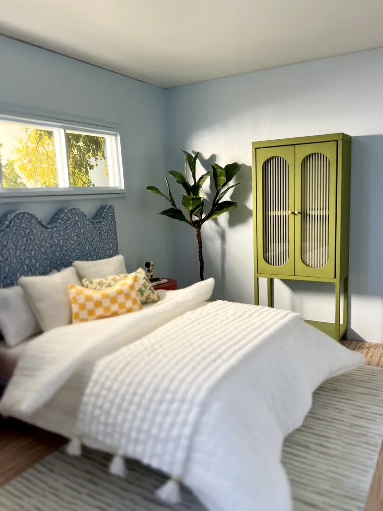 A cozy bedroom featuring a bed with a blue, patterned headboard, white bedding, and decorative pillows. A small window with view of trees, a side table with a lamp and clock, a tall indoor plant, and a bright green cabinet against a light blue wall.