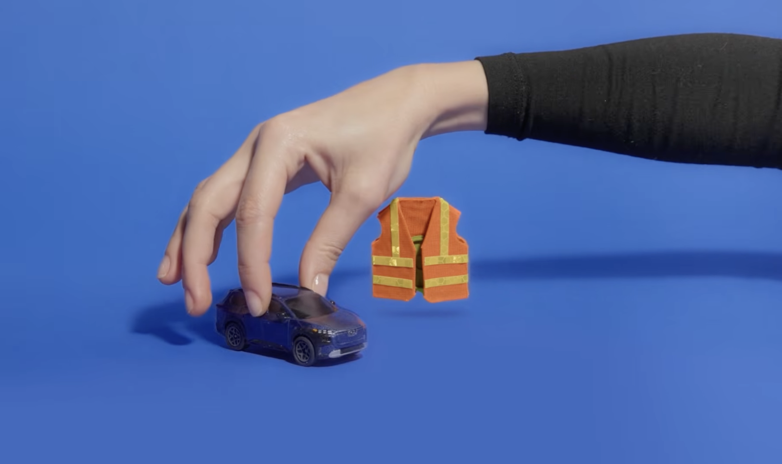 A hand with a black sleeve is reaching for a small toy blue car on a blue surface. Above the car, there is a miniature orange safety vest with yellow reflective stripes. The background is solid blue.