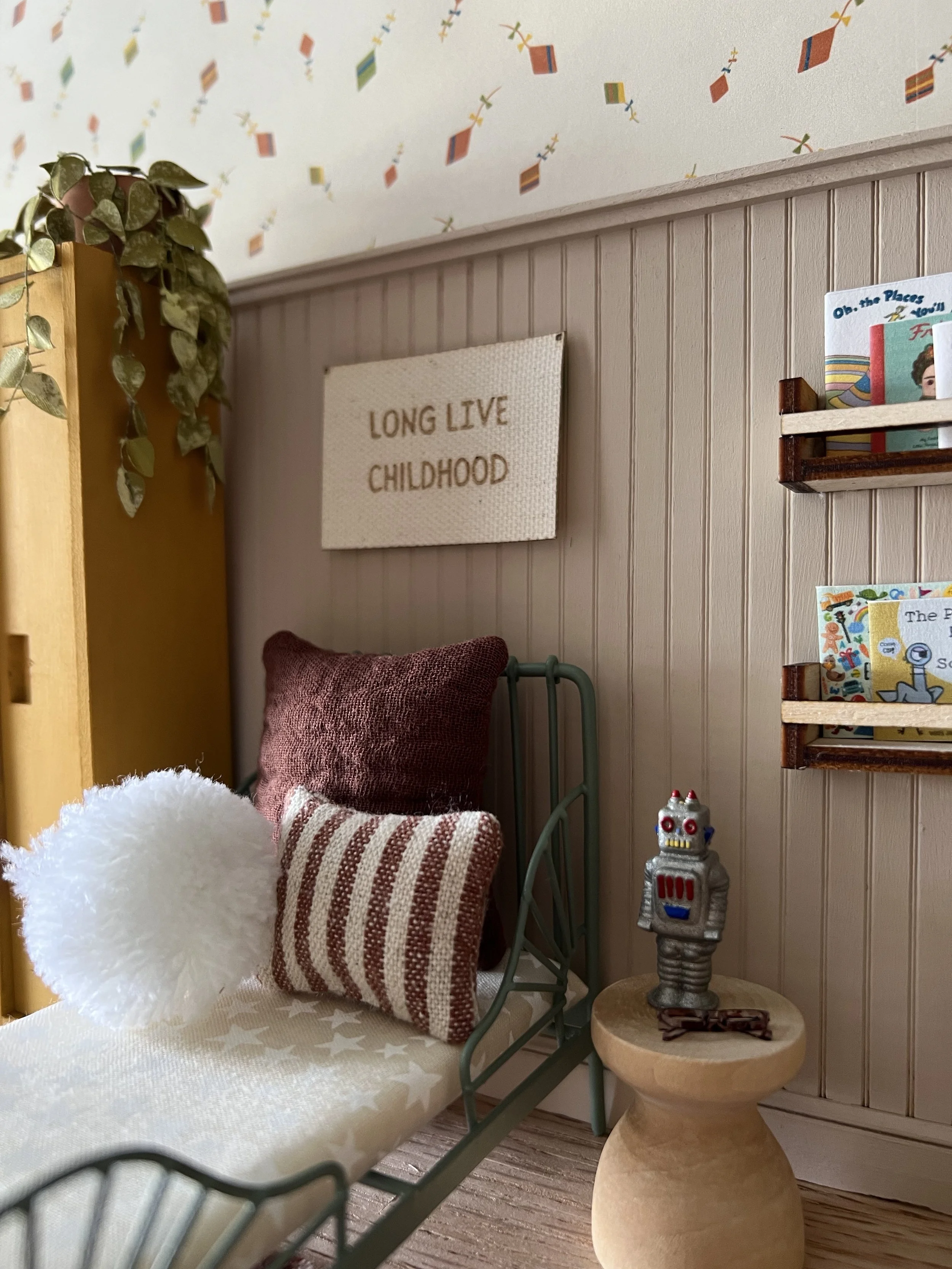 A cozy bedroom corner with a bed, red-striped pillow, and plush gray blanket, a beige beaded wall hanging reading 'Long Live Childhood,' a wooden shelf with colorful children's books, and a small robot figurine on a round wooden side table next to a 