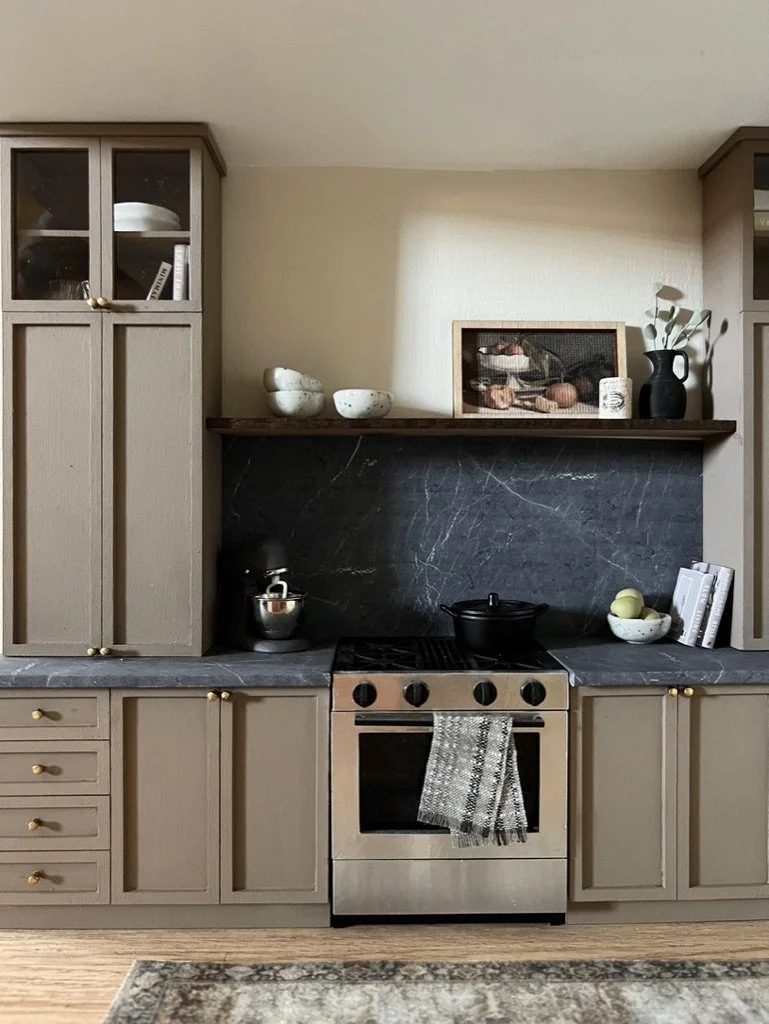 Kitchen with beige cabinets, black marble backsplash, stove with a gray and white towel, white bowls on shelf, framed nature scene, black vase with green leaves, and a countertop with books and fruit.