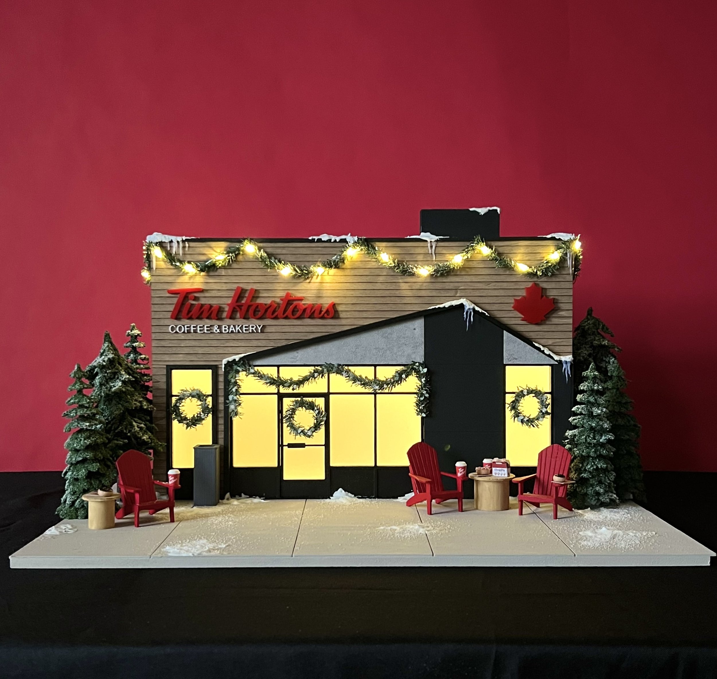 A festive storefront display with a red background, decorated with holiday lights, garland, snow, and Christmas trees. The store's sign reads "Tim Hortons Coffee & Bakery" and features a red maple leaf. In front, there are red chairs, a small table with holiday decorations, and red wrapping paper, creating a cozy holiday scene.