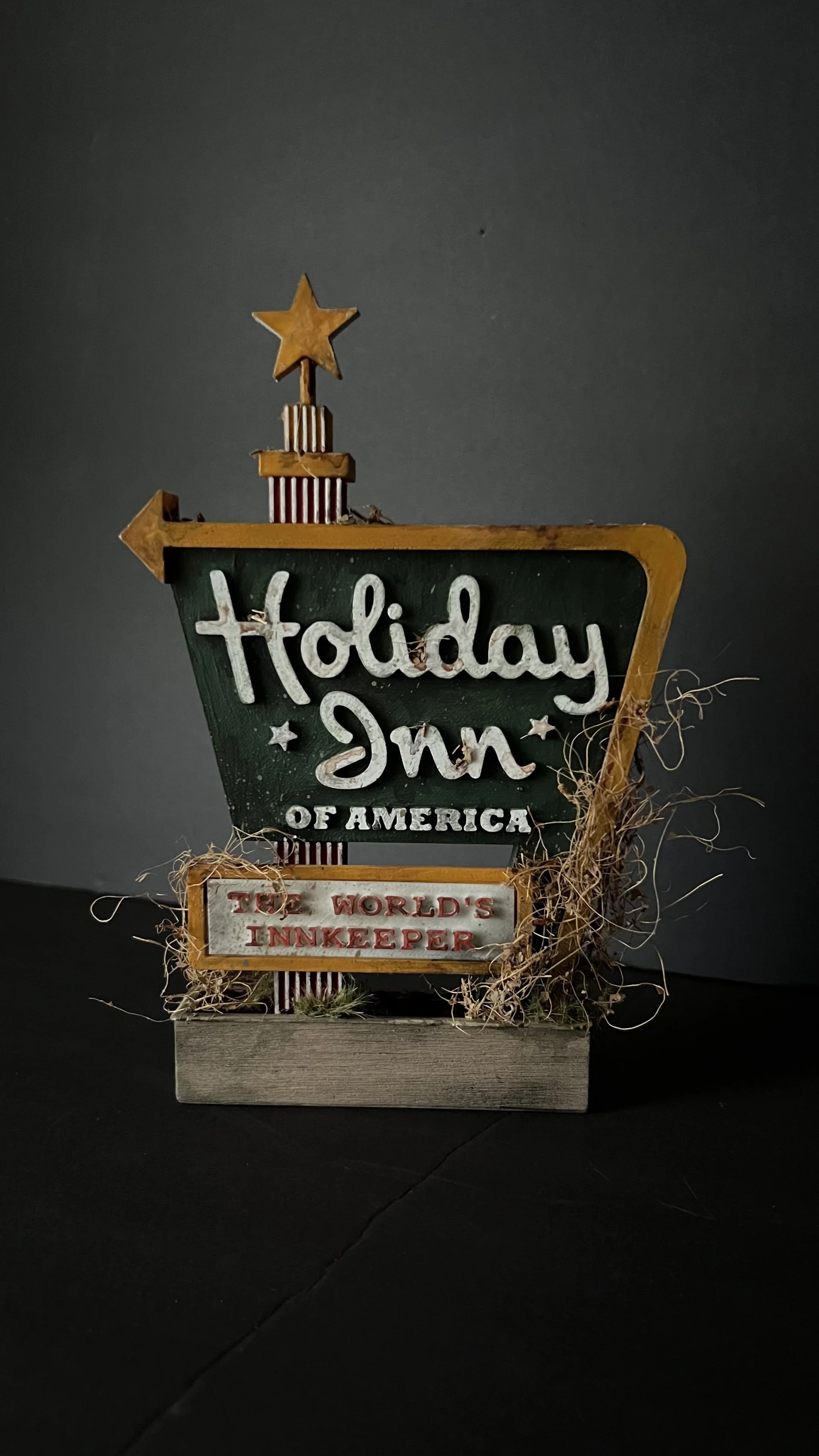 A decorative sign with a star on top reads 'Holiday Inn of America, The World's Innkeeper', with some decorative elements like stars and striped patterns, and is placed in a small planter with moss and twigs.