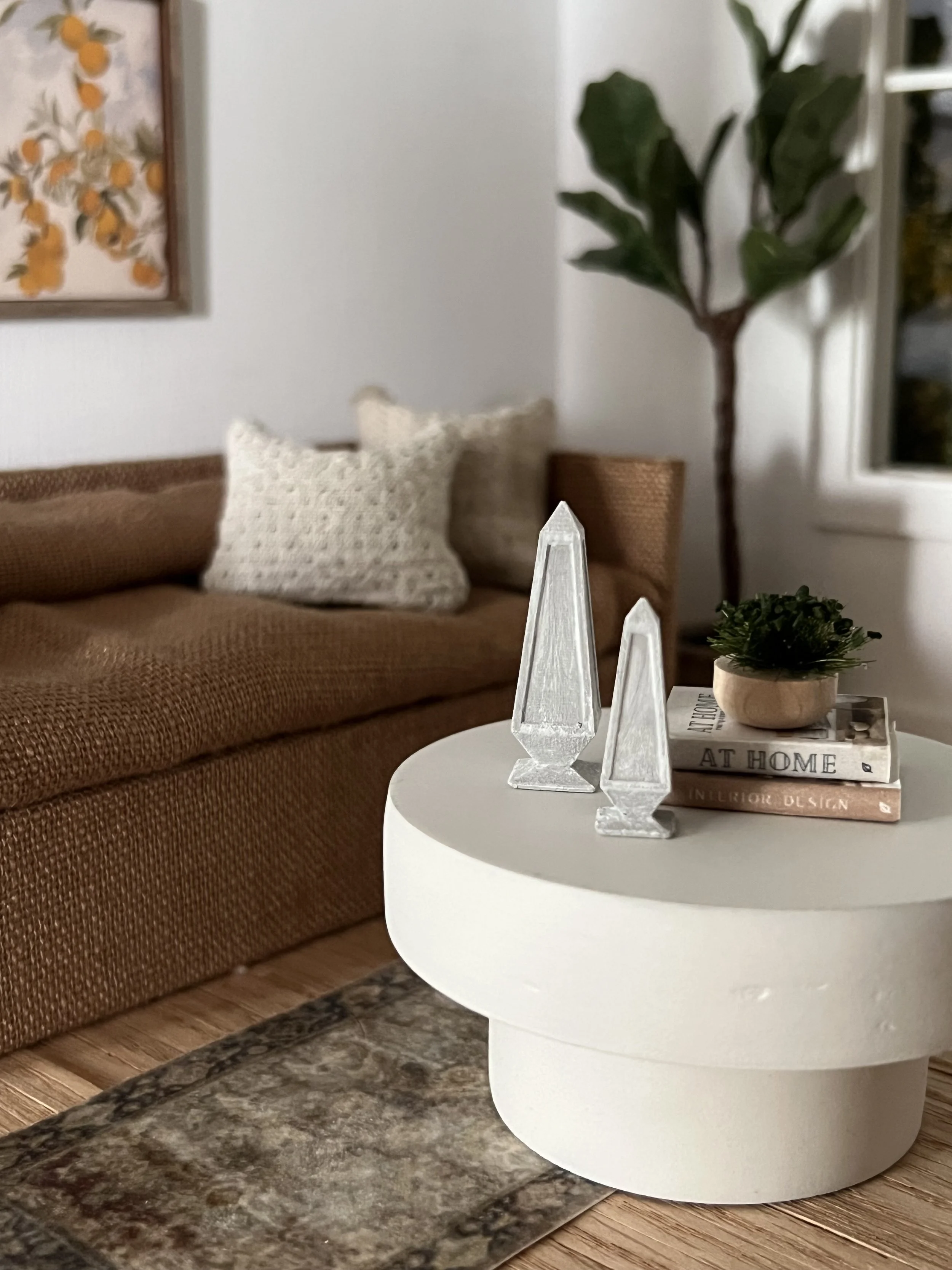 Close-up of a white round side table with decorative items on top, including a small green plant in a wooden pot and two white geometric crystal-like objects. In the background, there's a brown sofa with a textured fabric, beige and brown throw pillo