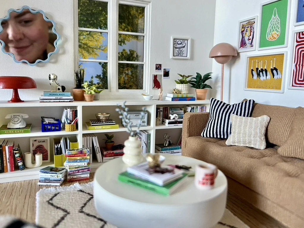 Cozy living room with a brown sofa, two striped pillows, and a white pillow, a white bookshelf filled with books and decor, a round white coffee table with various items, artwork on the walls, and a window showing trees outside.