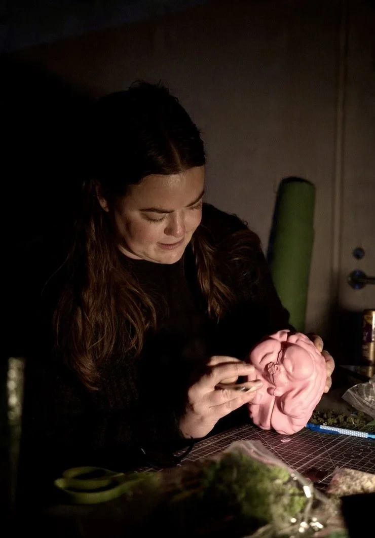 Woman sculpture artist carving a pink clay sculpture of a baby in a dimly lit studio.