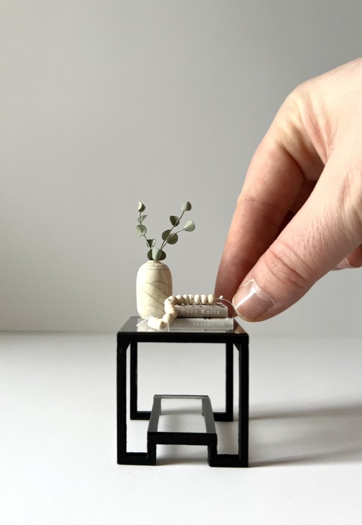 Miniature black table with a beige vase holding green leafy branches, a small book with a string of white beads, and a person's hand adjusting the beads.