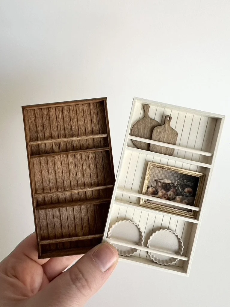 Two miniature wooden shelves, one in brown and one in white, each with two horizontal racks and decorative items, held by a person's hand against a plain wall.