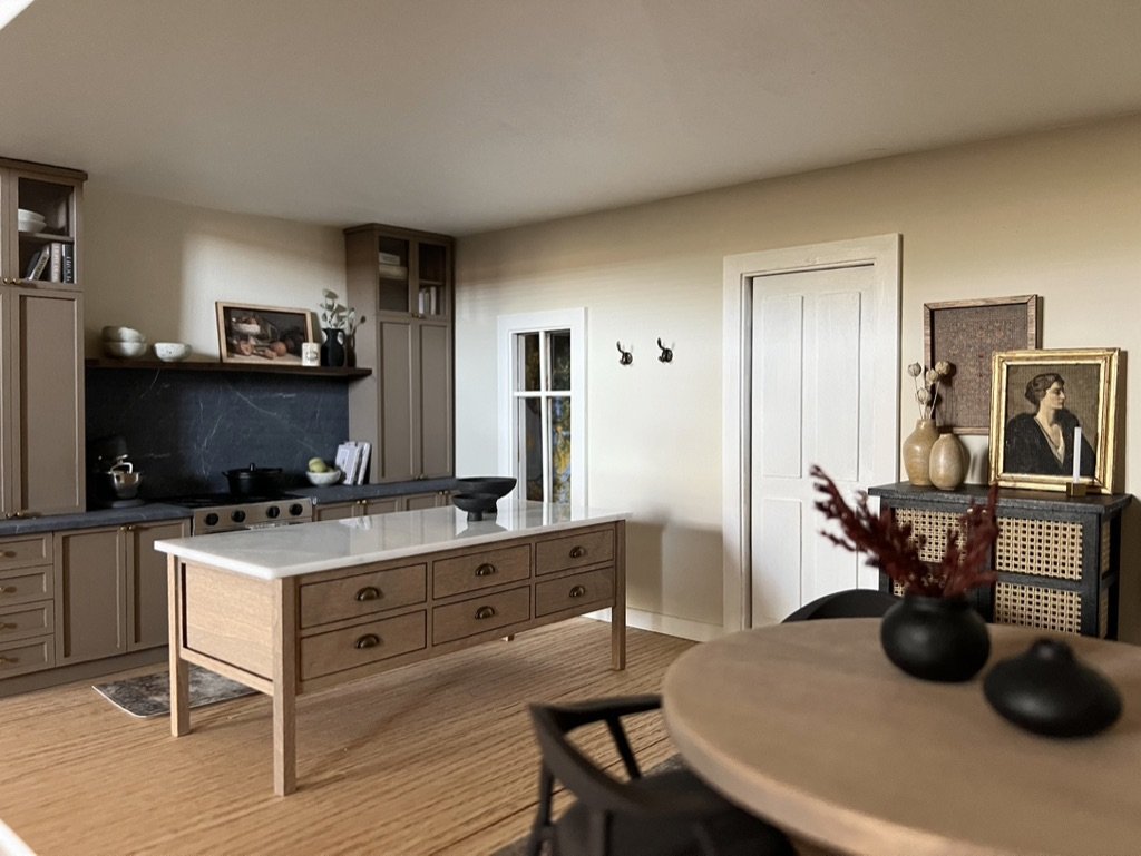 The image shows a cozy, well-decorated kitchen with beige walls, a white door, and light wood flooring. There is a kitchen island with drawers and a black bowl on top. Along the wall, there are gray cabinets with open shelves and a black backsplash. 