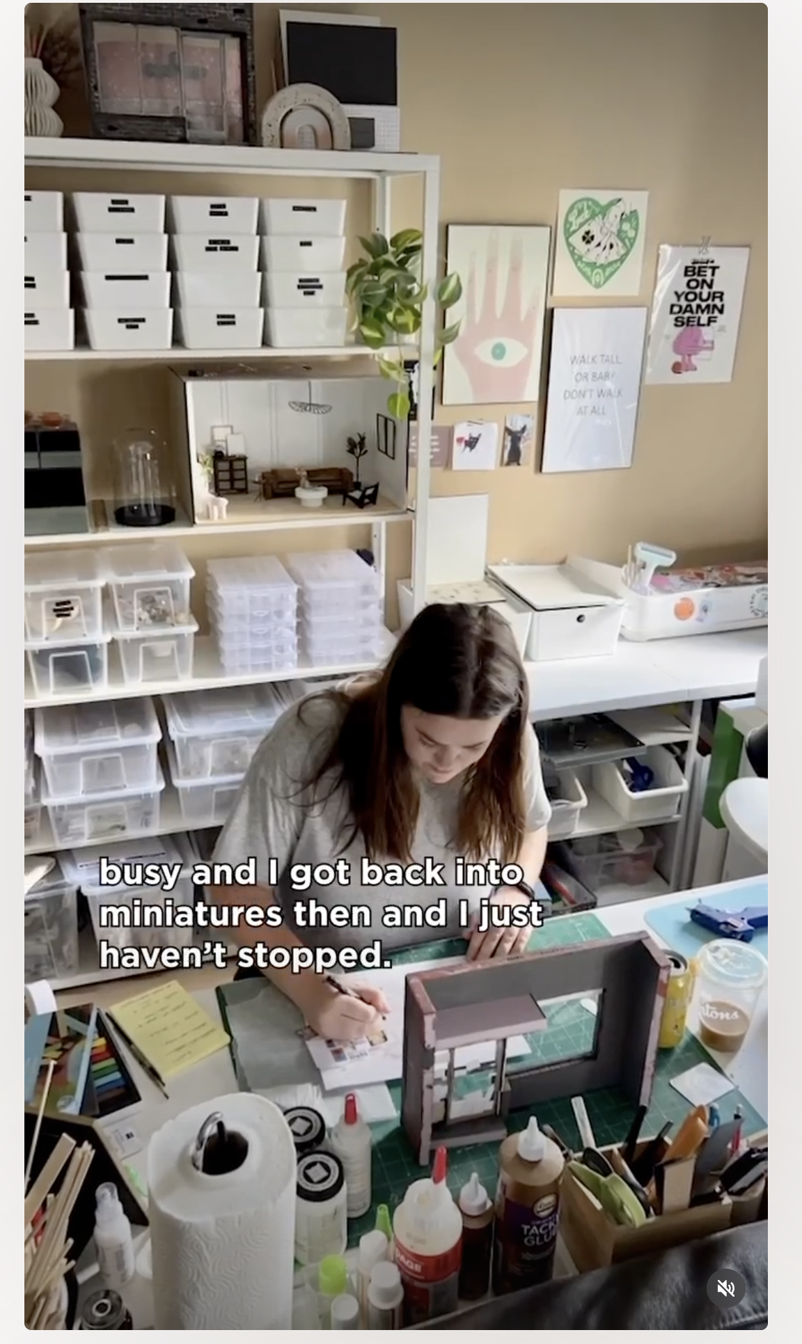 A woman working at a craft or miniatures table in a workshop surrounded by art supplies, miniature models, and storage bins, with wall art and motivational posters in the background.