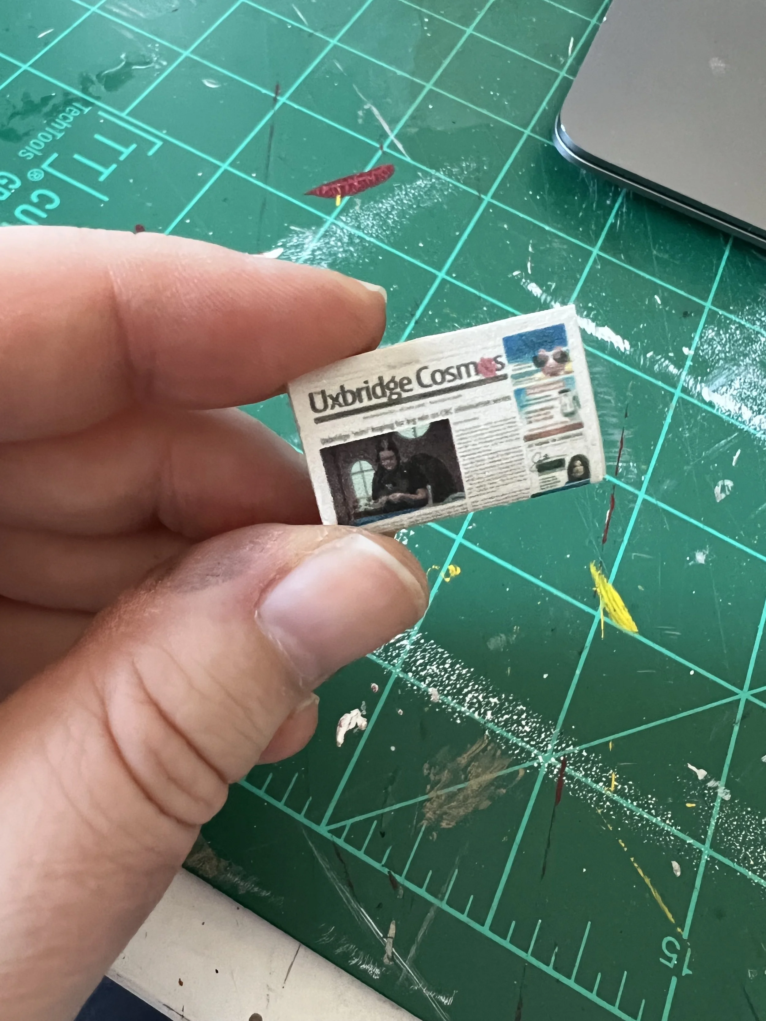 Close-up of a hand holding a small printed newspaper or magazine titled 'Uxbridge Cosmos' against a green cutting mat with measuring grid lines. The background includes a silver laptop and a transparent plastic sheet.