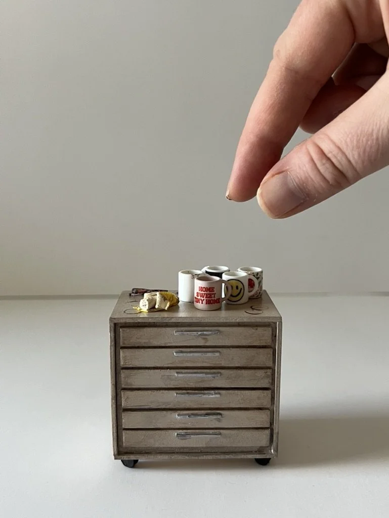 Miniature wooden cabinet with several coffee mugs on top and a tiny piece of sandwich, with a hand dropping a small object into the mugs.
