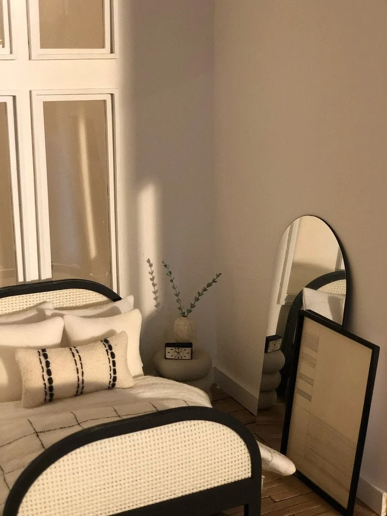 A bedroom with a bed, pillows, and a blanket, a small side table with a clock and vase with tall green branches, a large oval mirror leaning against the wall, and a framed picture or document on the floor.