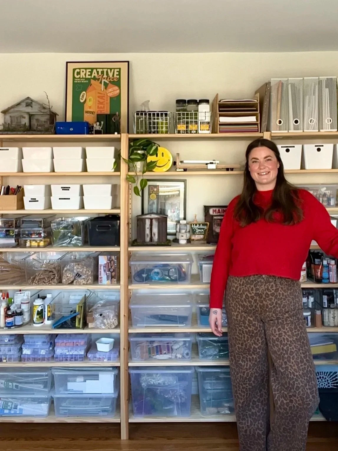 A woman wearing a red sweater and leopard print pants standing in front of organized shelves filled with storage bins, boxes, art supplies, and decorations.