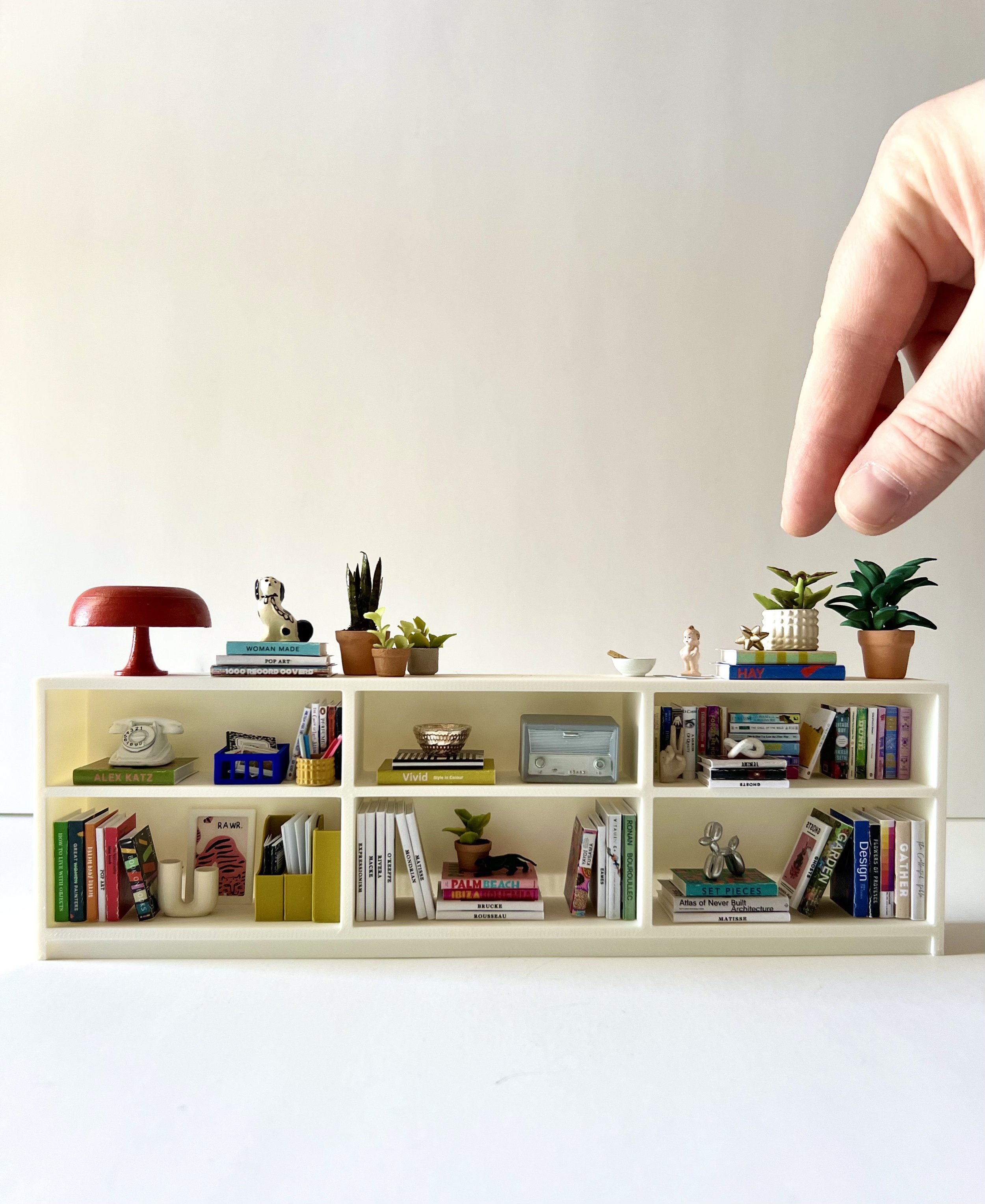 A white bookshelf filled with various books, small potted plants, decorative objects, and art pieces, set against a plain white wall. On top of the bookshelf are small plants, books, and decorative items. The background features a plain white wall.