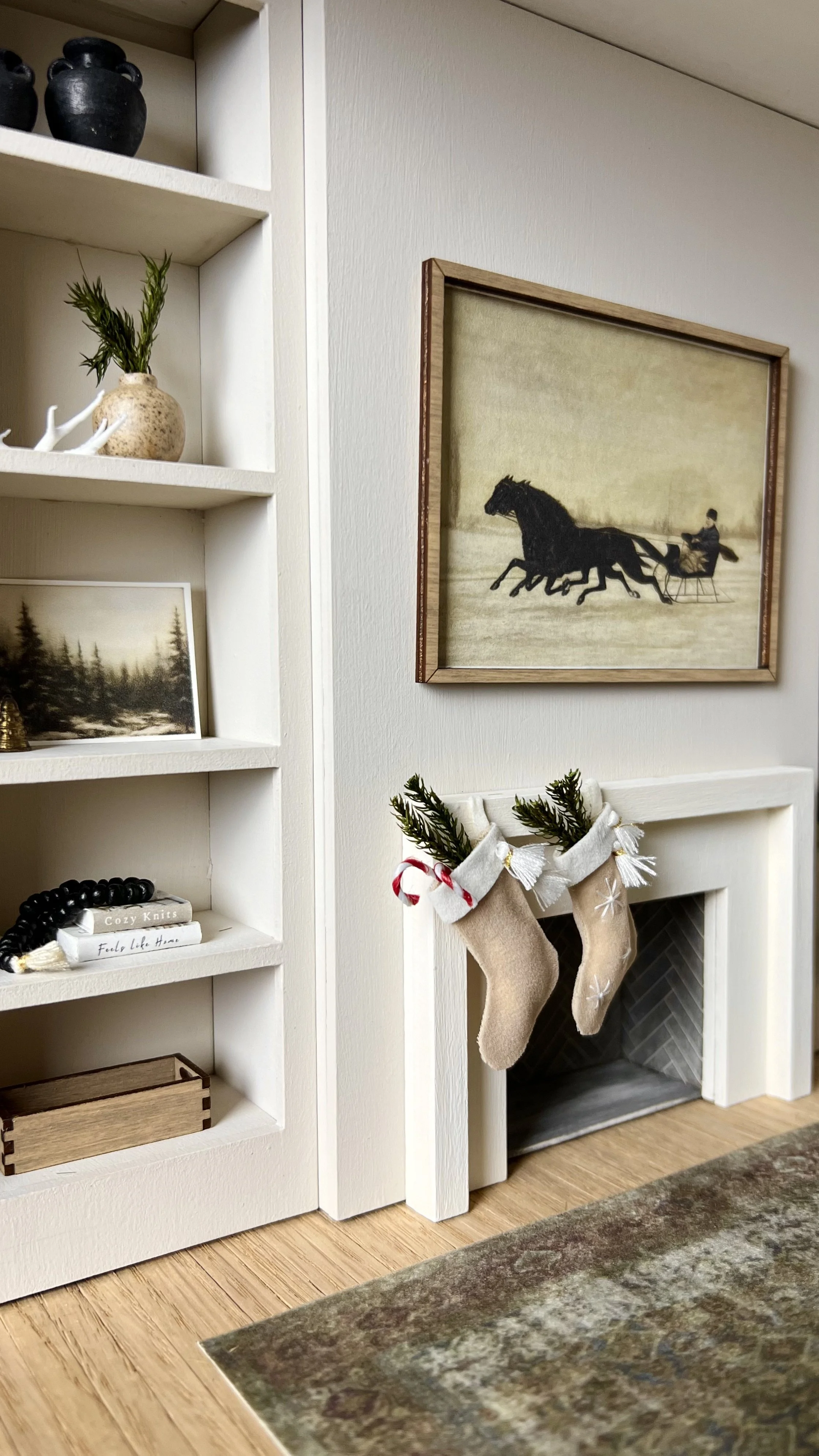 A white built-in bookshelf with decorative items including black vases, a small potted plant, a framed picture of a forest, a stack of books, and a beaded bracelet. Next to the bookshelf is a fireplace with two Christmas stockings hanging, and above 
