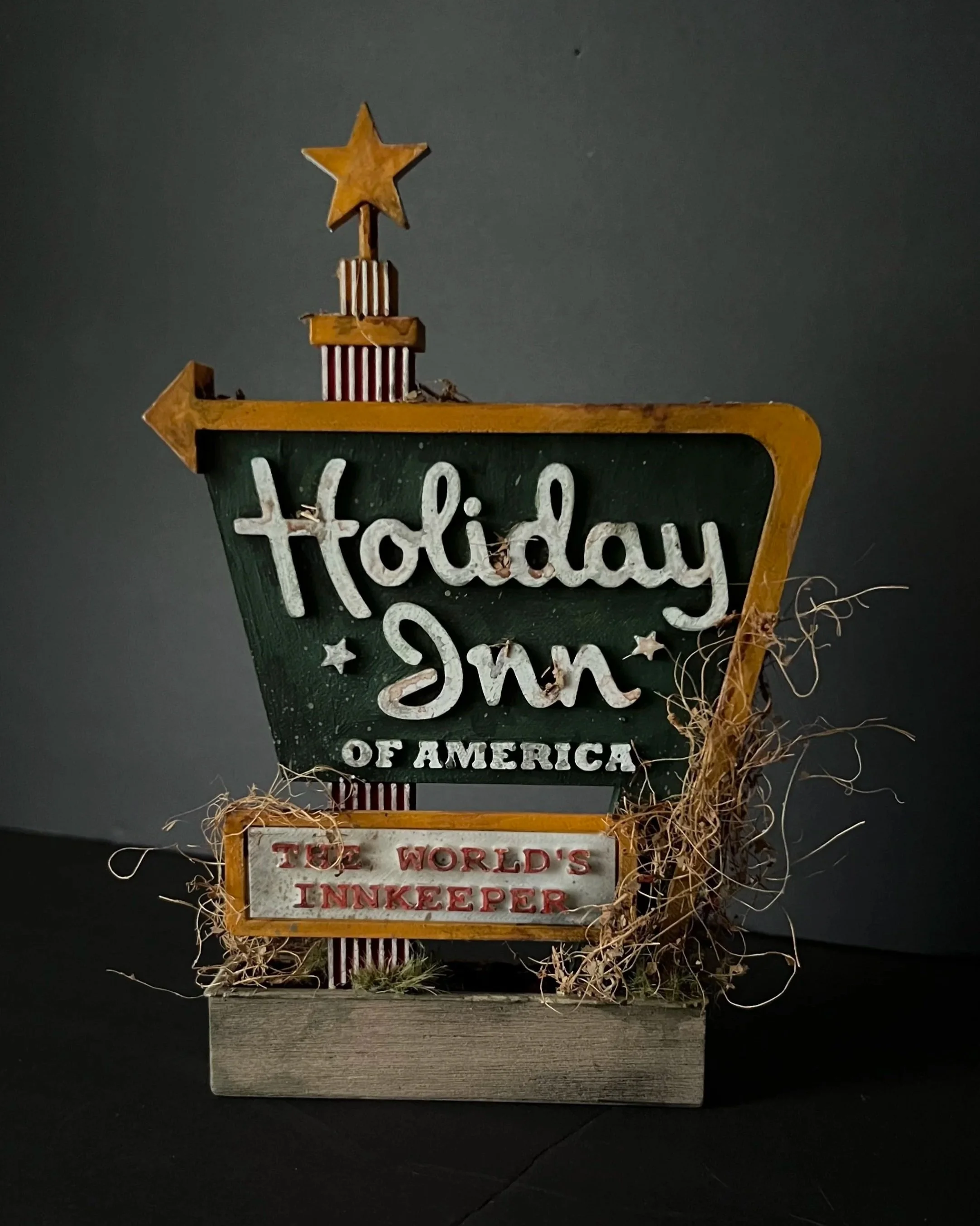 Decorative sign with a yellow star on top, featuring the words "today's" and "of America" in white, and a smaller sign below reading "the world's innkeeper" in red, surrounded by dried grass and leaves, against a dark background.