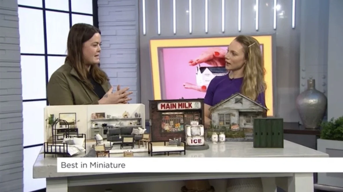Two women are standing at a table with miniature furniture and buildings, engaged in conversation. Behind them is a television screen displaying a pink background with a hand holding a phone. The table displays a variety of miniature items including a house, a convertible, and a small milk stand labeled 'Main Milk' and 'Ice'.