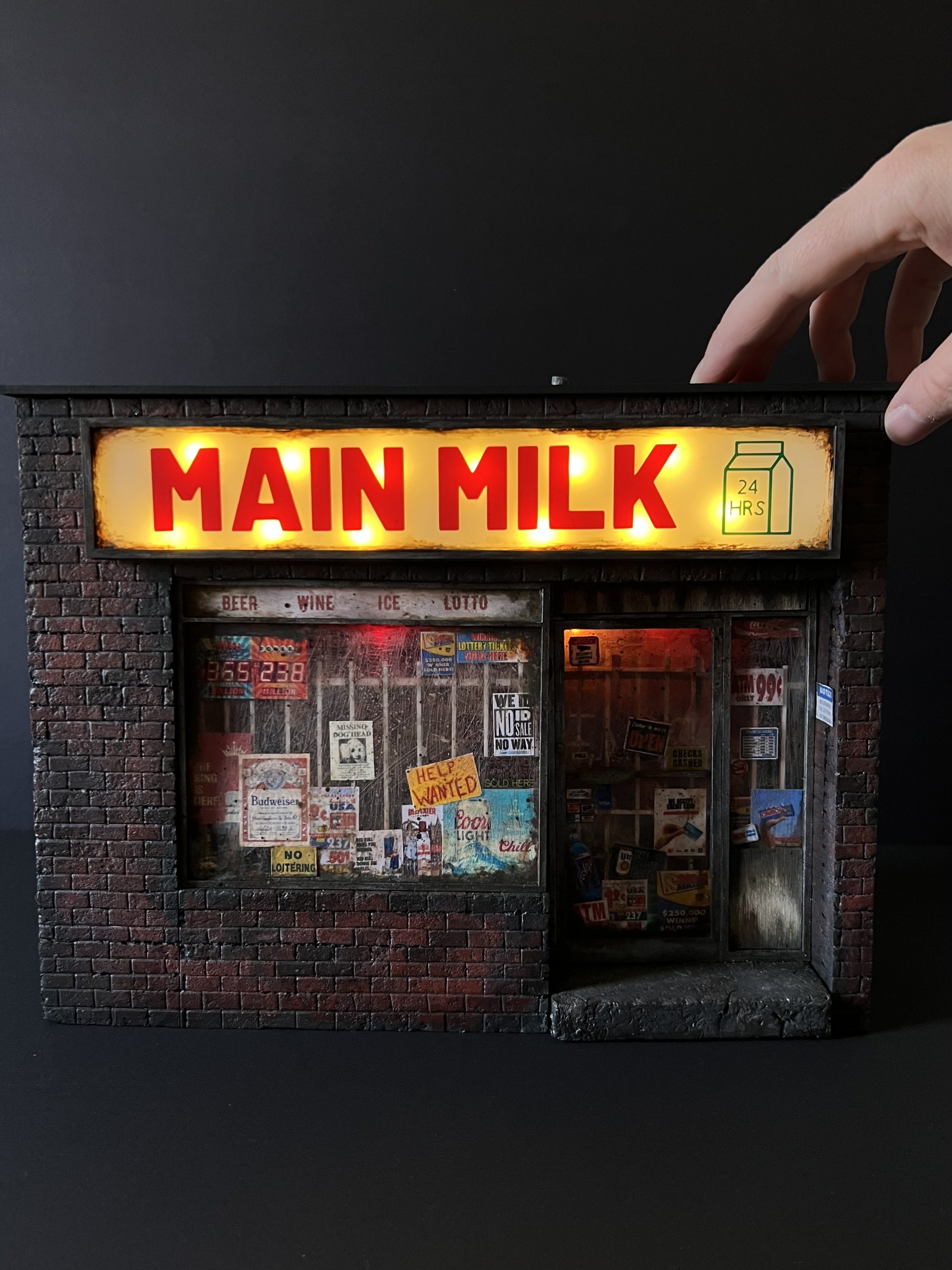 A miniature replica of a brick building with a sign that reads "MAIN MILK" illuminated with yellow lighting. The building resembles a small shop or vending machine with various posters, signs, and advertisements in the window, including notices for beer, wine, ice, lotto, and a handwritten sign that says "Help Wanted." A hand is visible holding the replica from the top right corner.