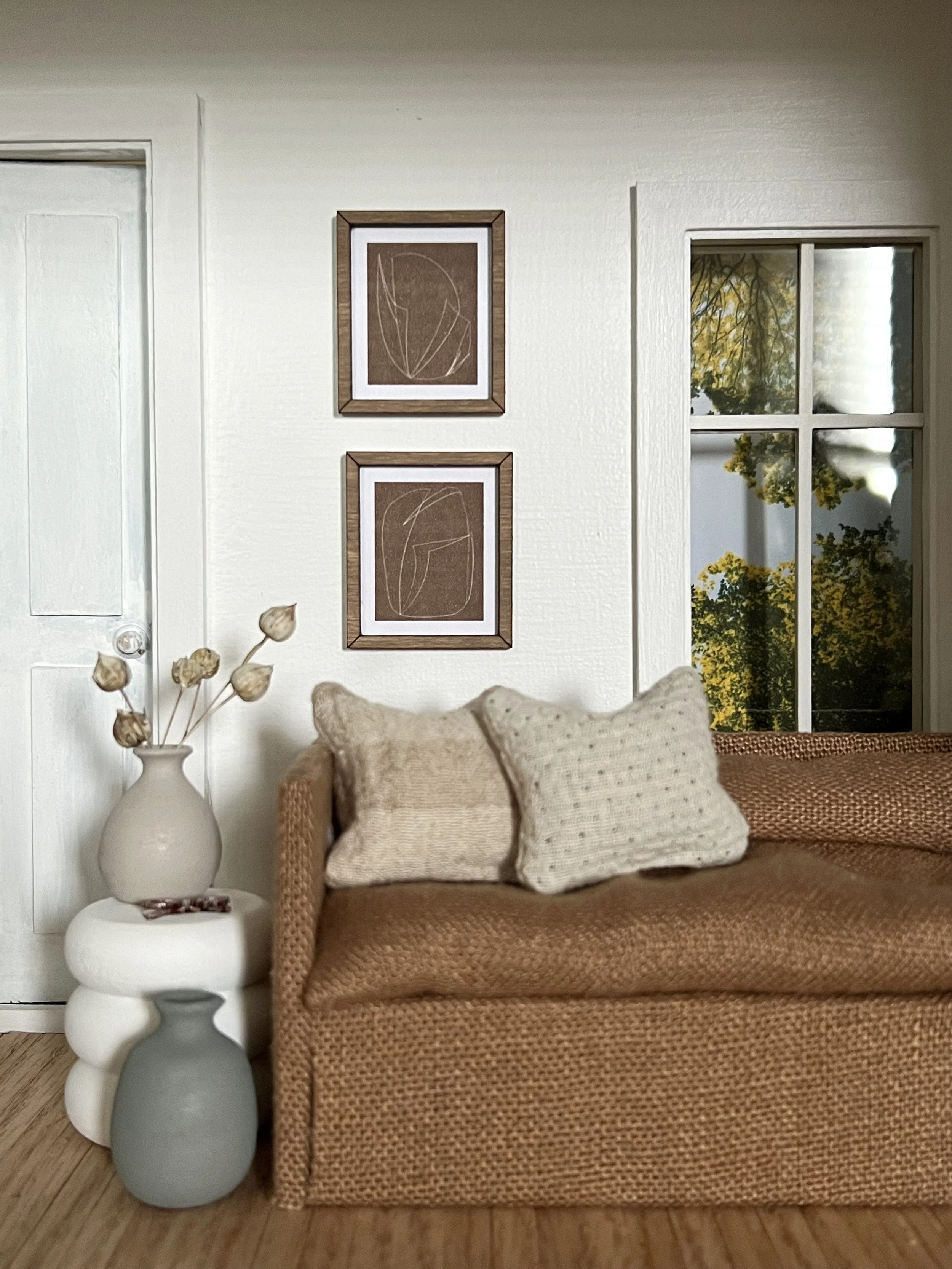 Living room corner with a wicker sofa, beige pillows, a white side table with tiered vases, a vase with dried flowers, framed art on the wall, and a window showing trees outside.