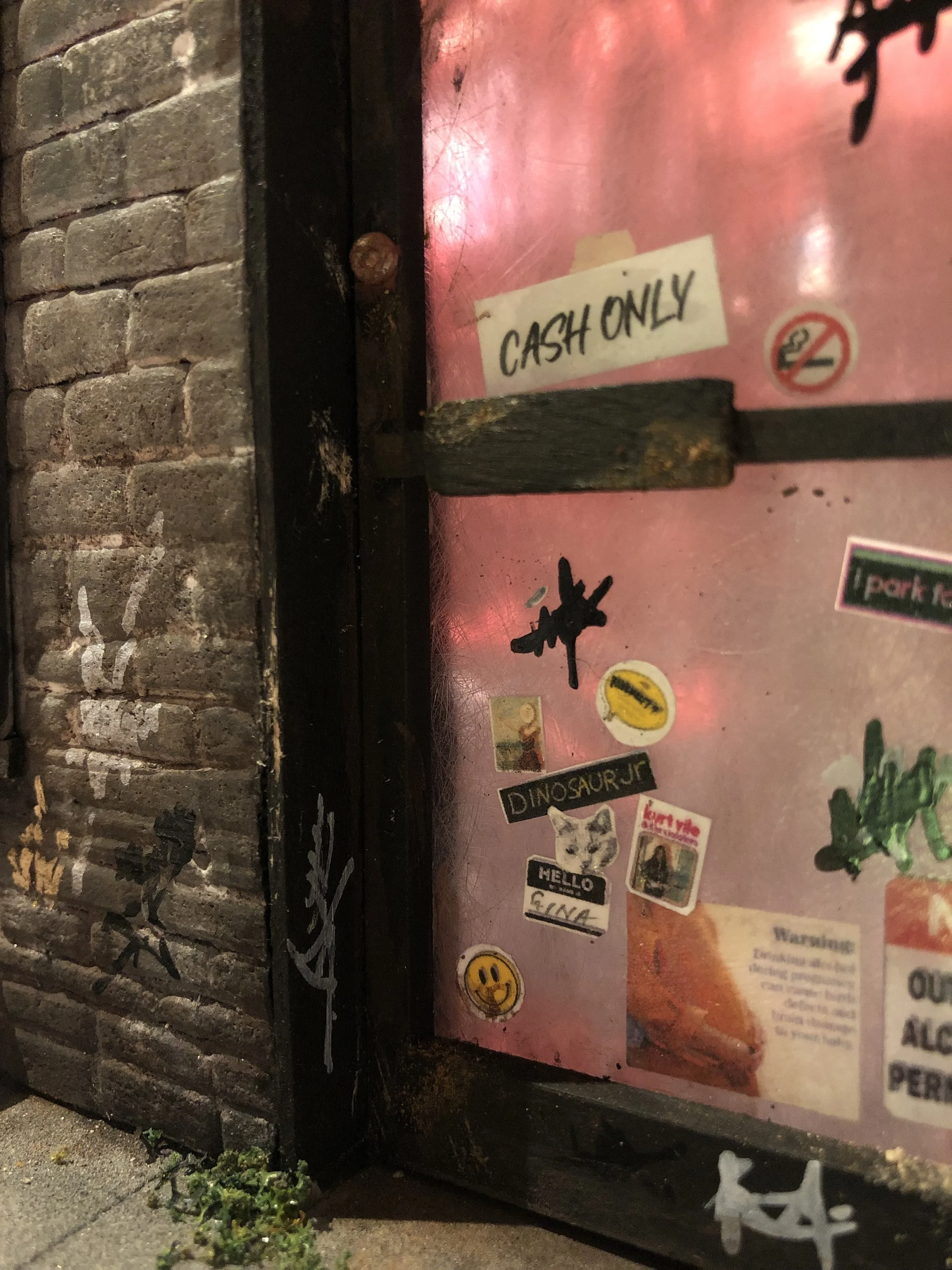 Close-up of a brick wall and a pink metal surface with various stickers and signs, including a yellow smiley face sticker and a sign that reads 'CASH ONLY'.