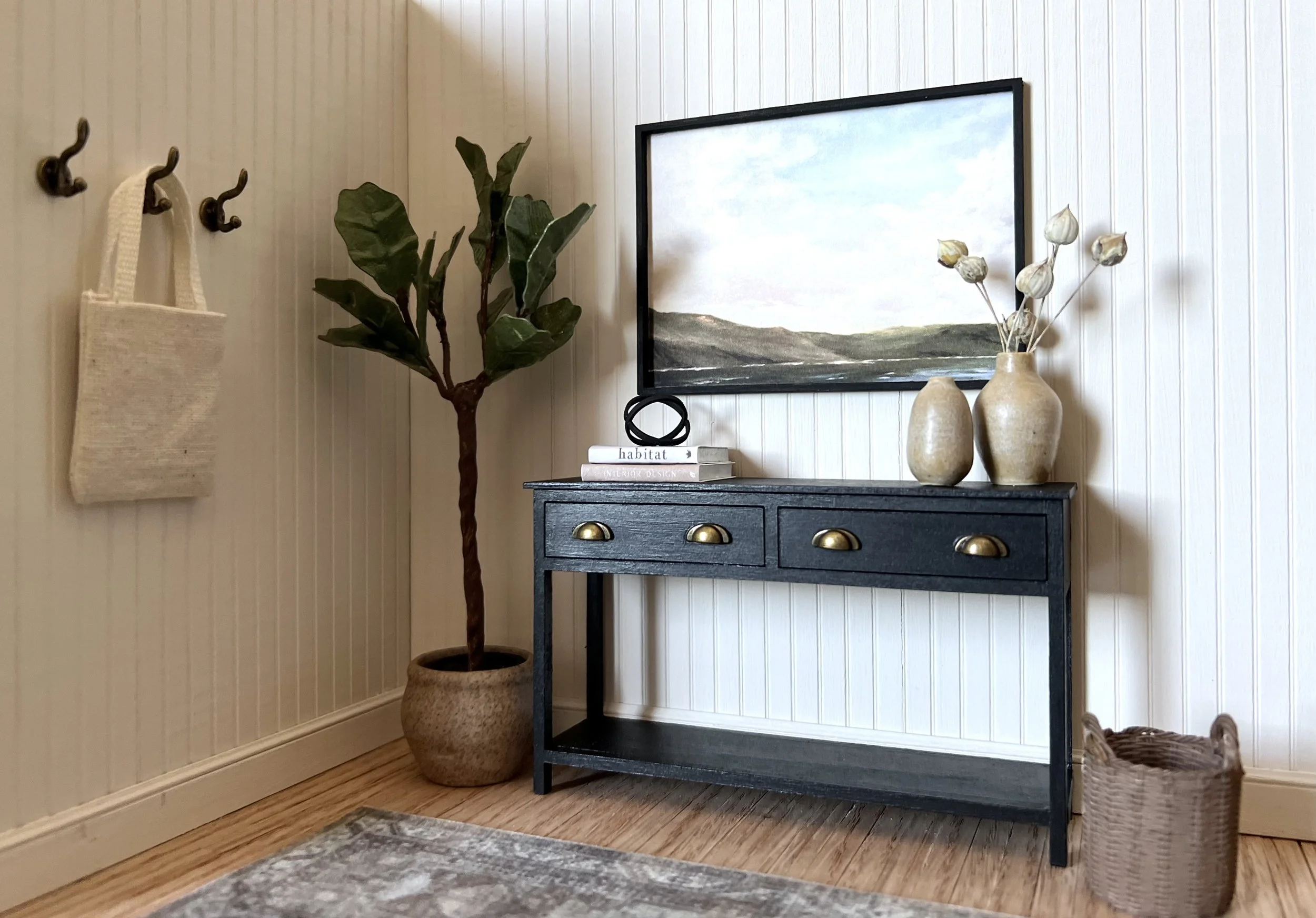A corner of a room with a black console table, large potted plant, decorative vase with dried flowers, fishing net wall decor, and framed landscape painting on cream-colored beadboard walls.