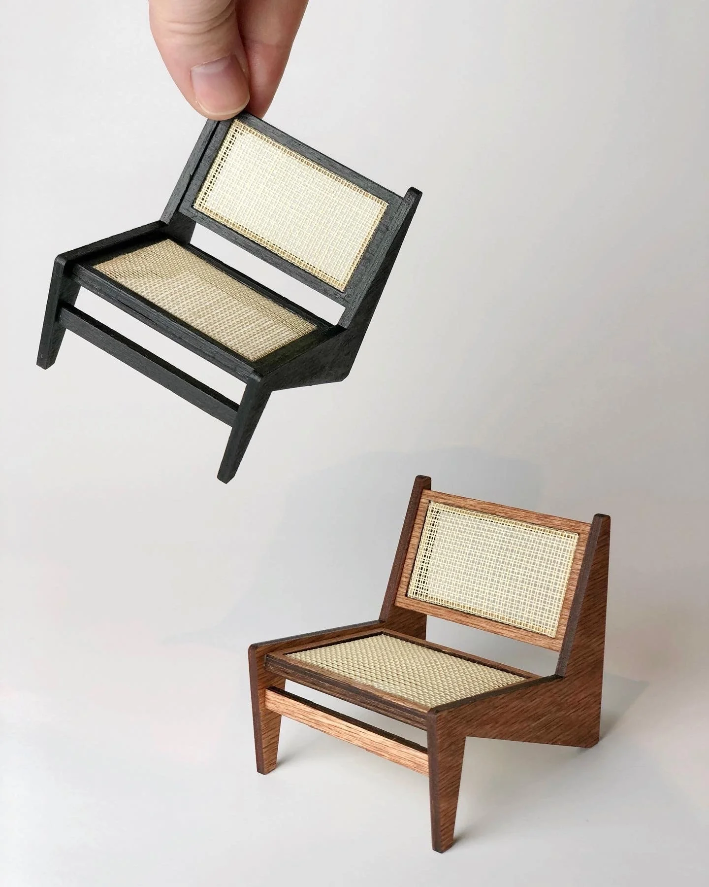 Close-up of a hand holding a miniature black wooden chair with a woven cane seat and backrest, with a similar brown wooden chair below, both set against a plain white background.