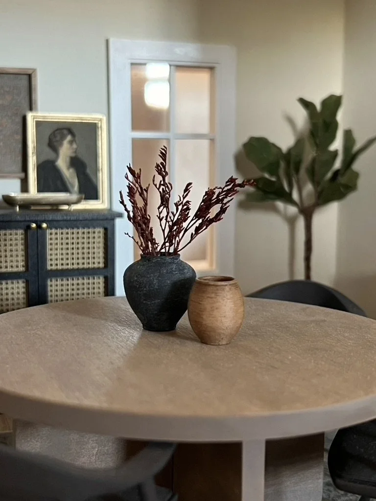 Decorative vases with dried branches on a wooden table in a cozy living room with framed paintings, a window, a large green plant, and a rattan-front cabinet in the background.