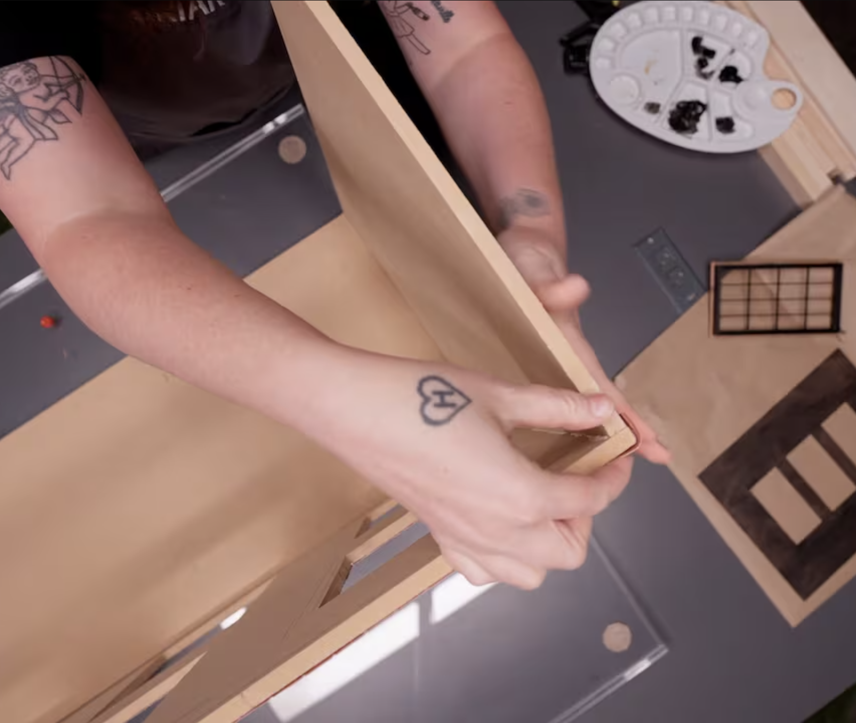 Person holding a wooden puzzle piece, with a palette of black paint and a small grid frame on a wooden surface nearby.