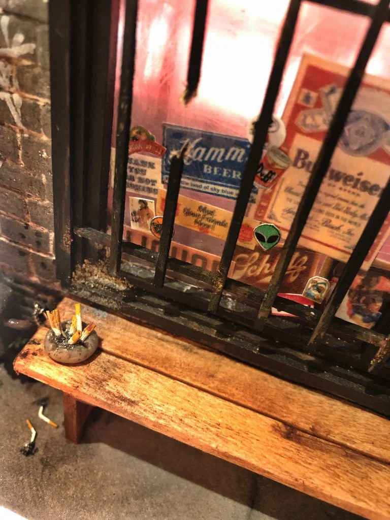 View through black iron bars showing a wooden bench with a small ashtray filled with cigarette butts in the foreground, and vintage beer advertisements on the pink wall behind the bars.