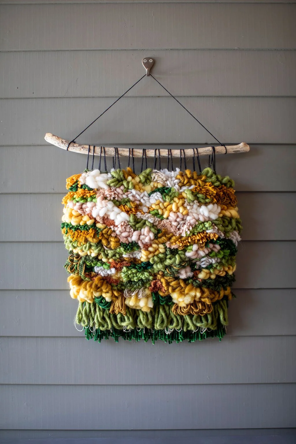 A colorful, textured wall hanging made from various yarns and fibers, suspended from a piece of driftwood attached to a hook on a gray wooden wall.