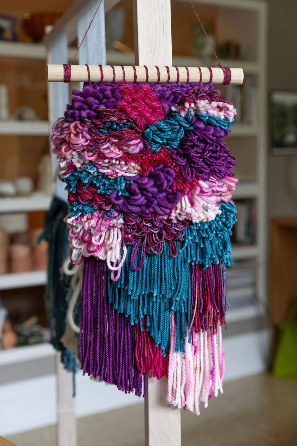 Colorful textured yarn wall hanging with purple, pink, blue, white, and red yarns on a wooden dowel.