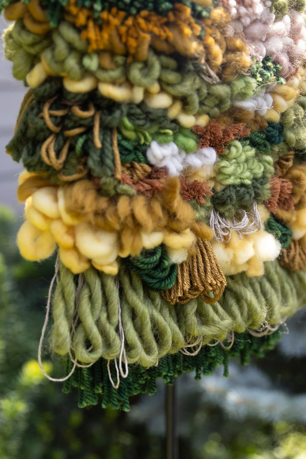Close-up of a colorful, textured yarn or wool wall hanging with various shades of green, yellow, brown, and white.